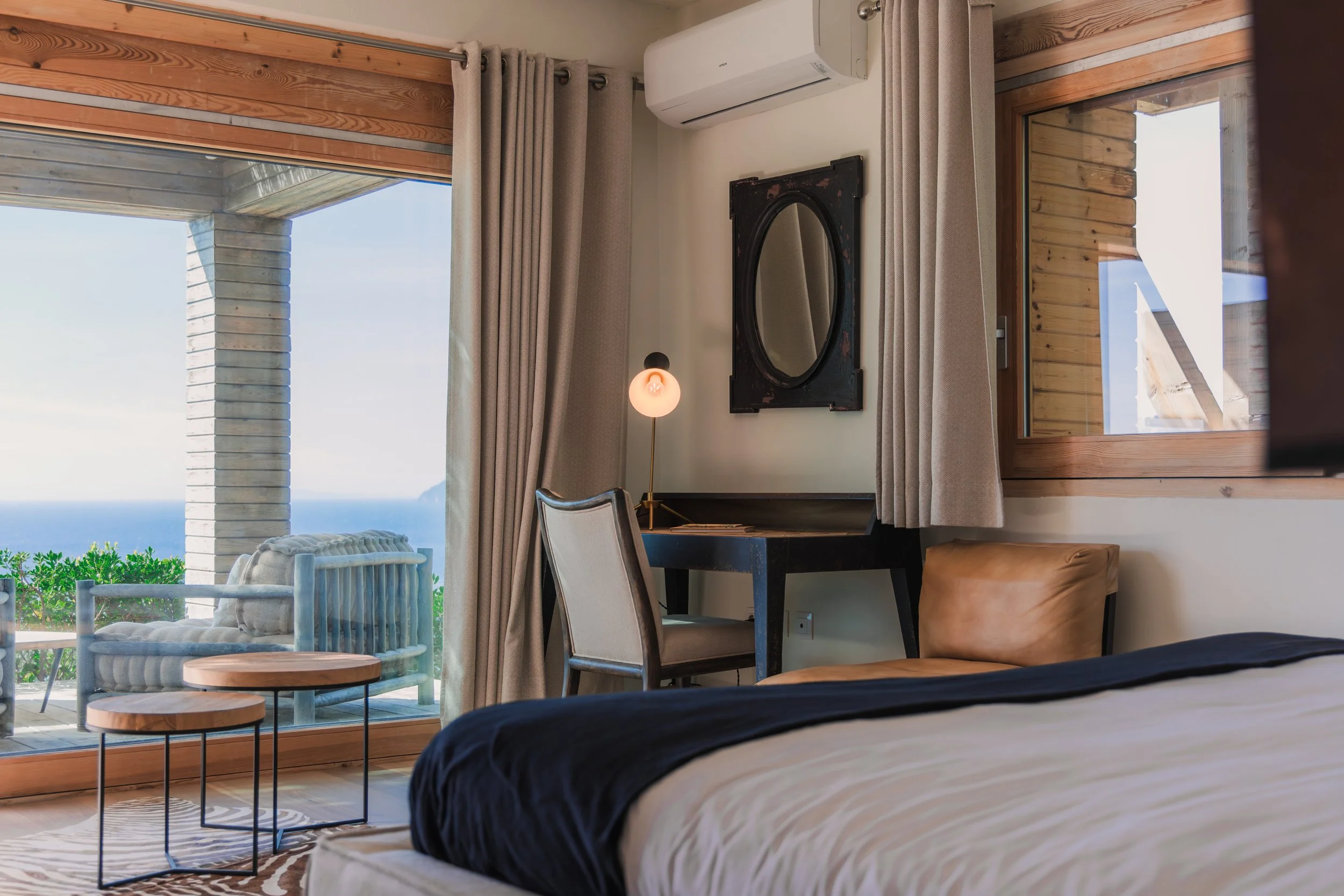 Hotel room with a bed, desk, chair, mirror, air conditioner, and large glass door showing an outdoor patio and ocean view.