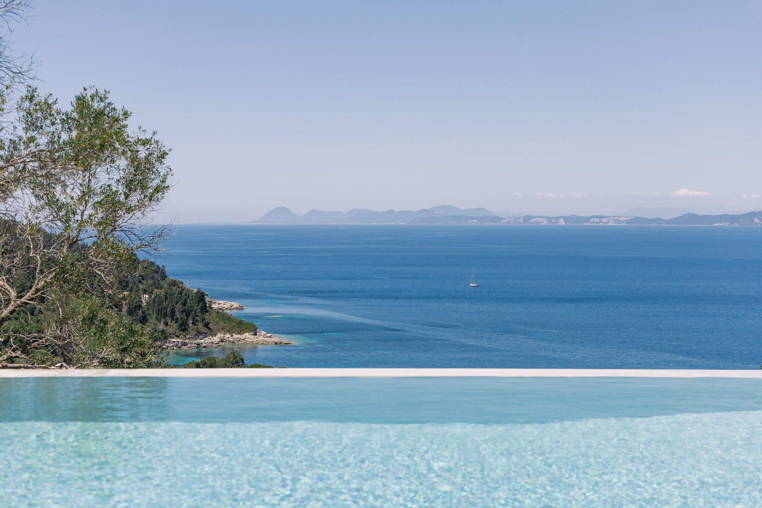 Scenic view of the ocean with a sailboat, distant mountains, and a clear blue sky, seen from an infinity pool edge.