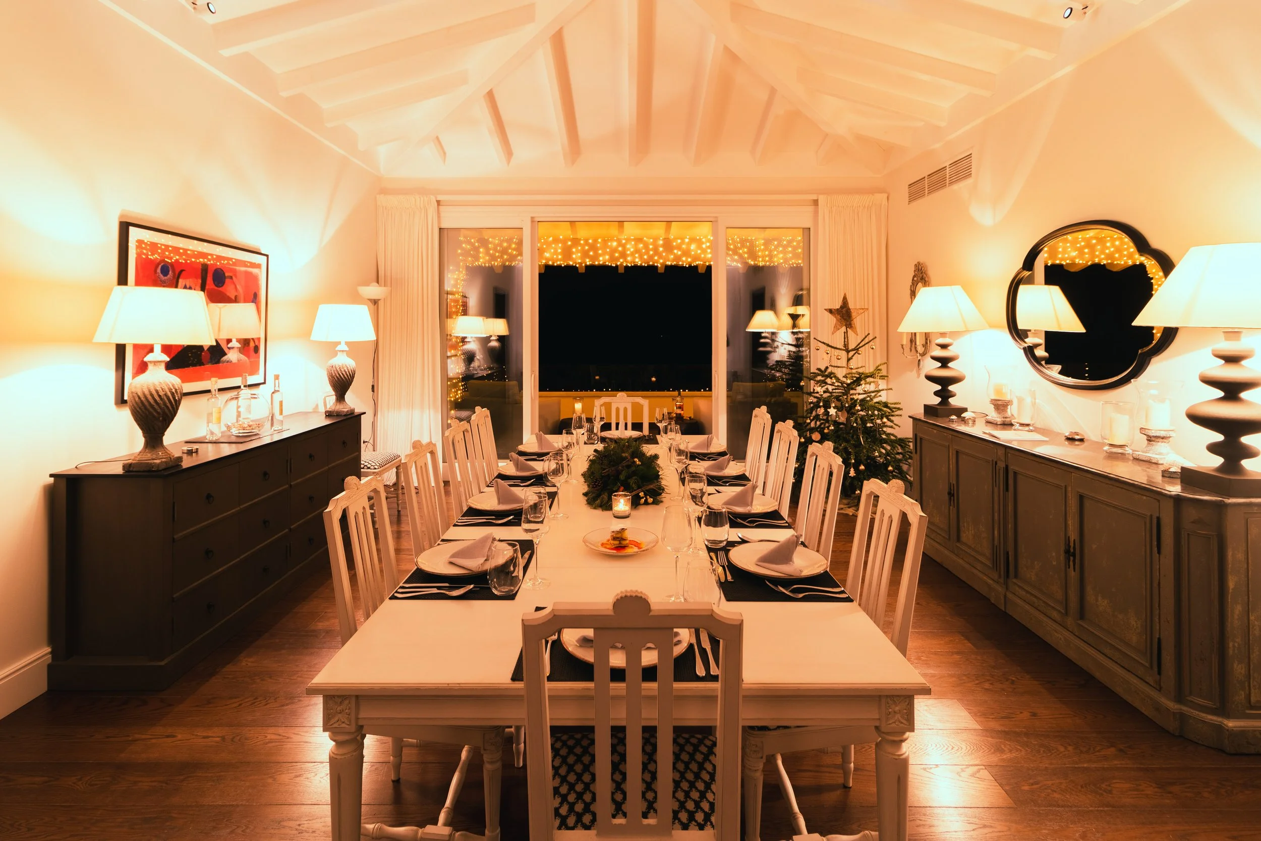 Christmas decorated dining room with table set for a meal, illuminated by warm lamps, with a small Christmas tree and fairy lights outside the window.