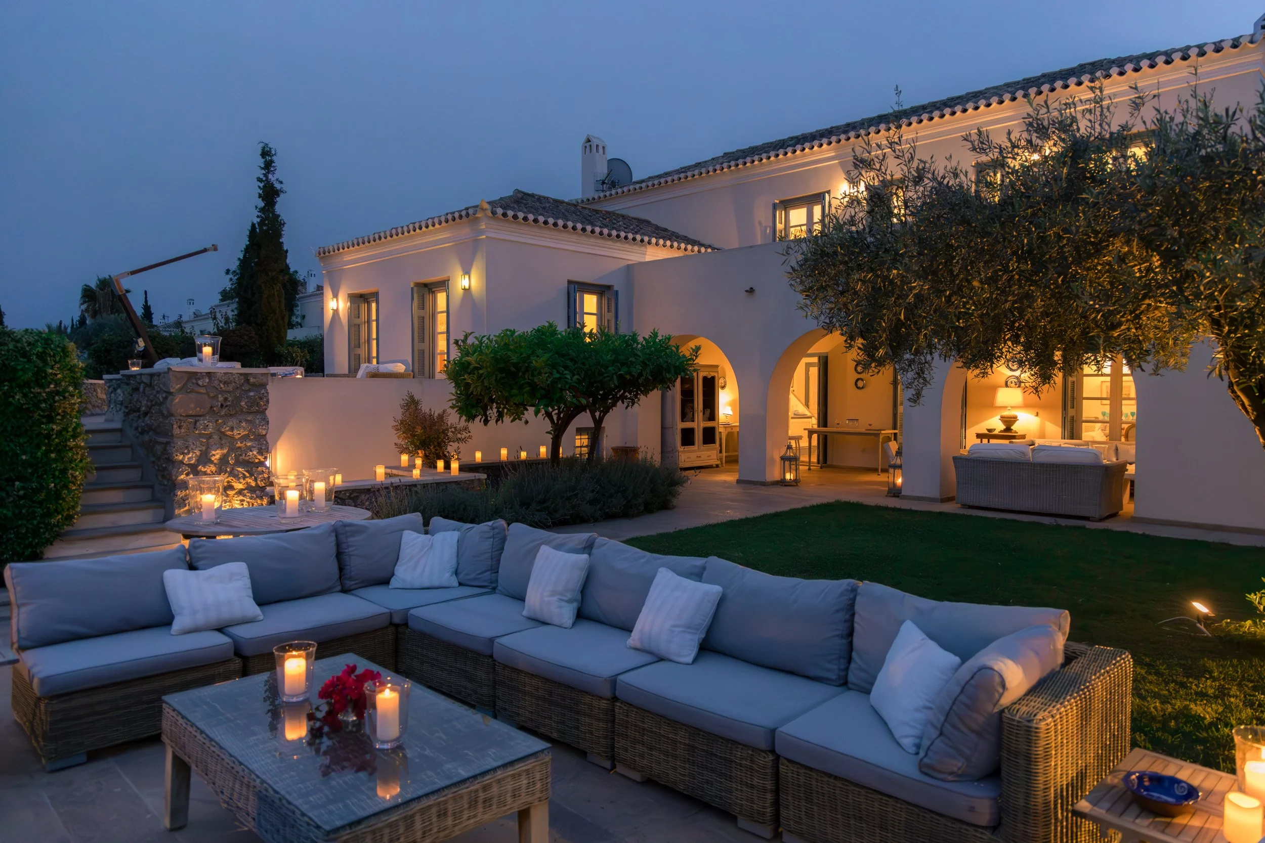 A cozy outdoor seating area at dusk with a large sectional sofa with white cushions, candles, and a glass-topped table surrounded by candles and flowers, facing a white Mediterranean-style villa with arched entryways and warm interior lighting.