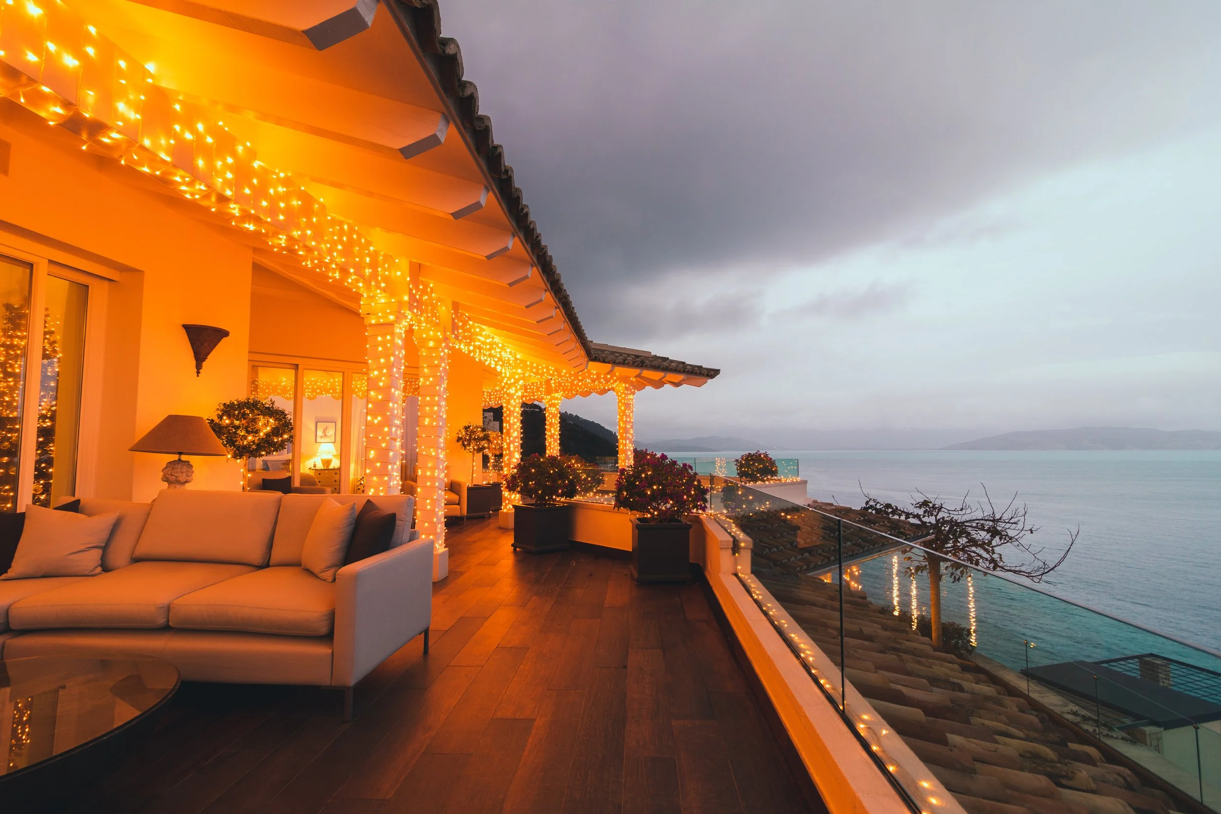 A spacious balcony decorated with warm yellow string lights, overlooking the ocean. It features a white sofa, potted plants, a lamp, and a view of a cloudy sky over the water.