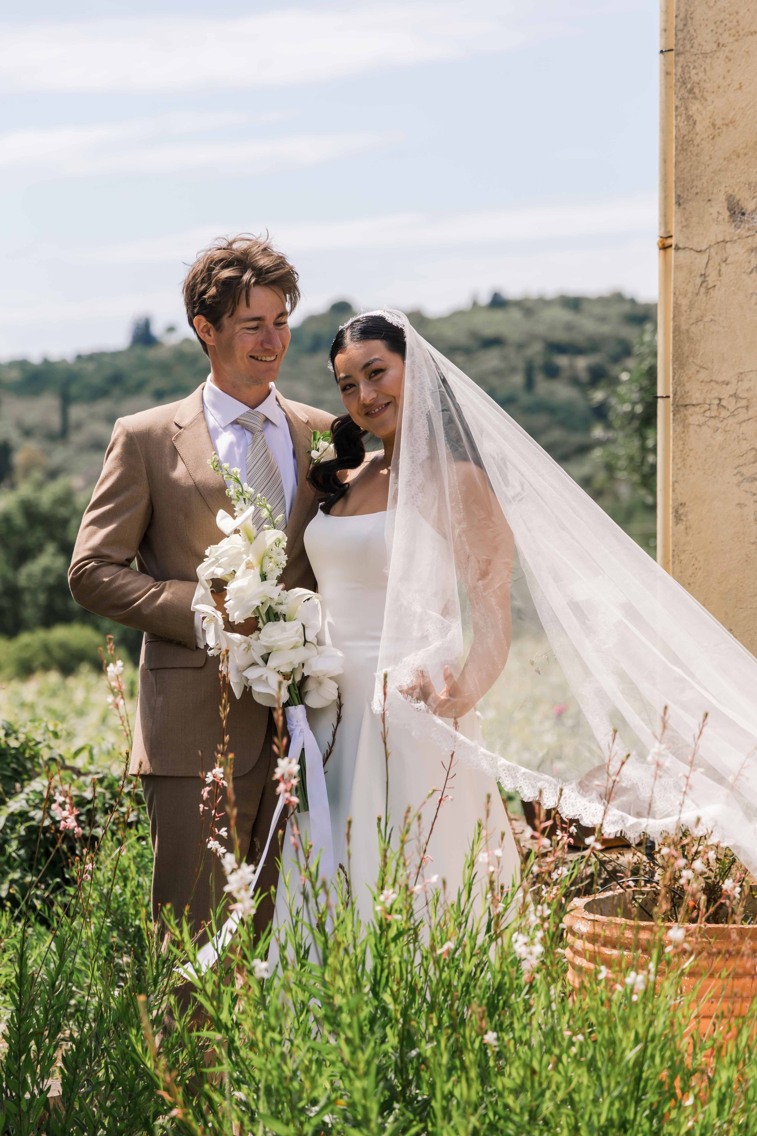 A wedding couple standing outside in a garden, with a scenic hillside in the background. The bride wears a white strapless gown and veil, holding a bouquet of white flowers. The groom wears a beige suit and tie, smiling at her.