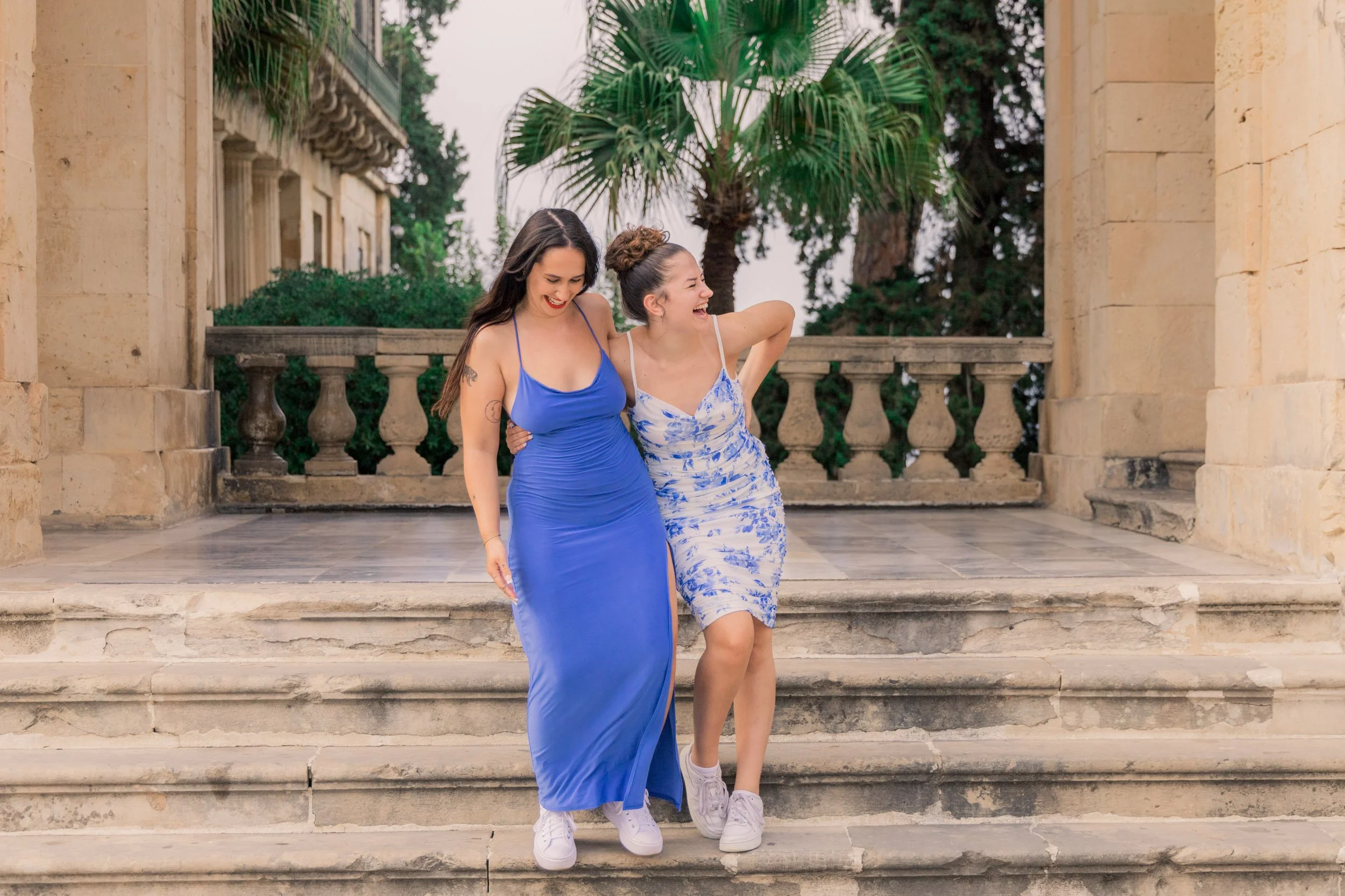 Two women in blue dresses smiling and laughing as they walk down stone steps outside a historic building with columns and a balustrade, with lush green palm trees in the background.