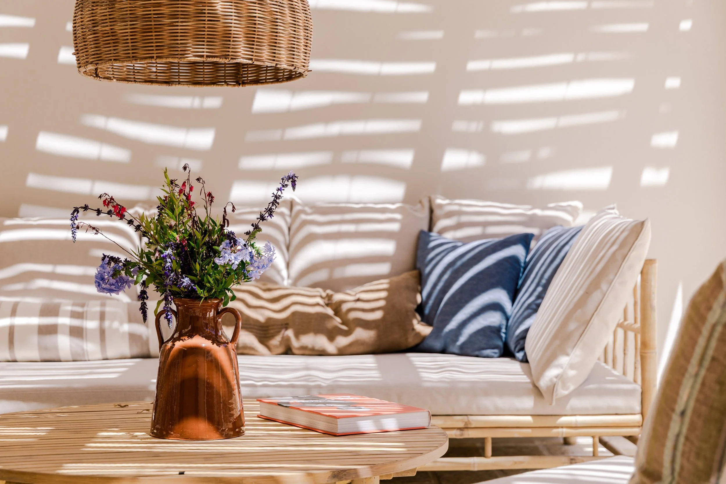 Sunlit outdoor patio with a wicker sofa decorated with striped and solid cushions, a wooden coffee table with a copper vase of purple and blue flowers, and a striped lounge chair partially visible on the side. Shadow patterns from a slatted overhead 