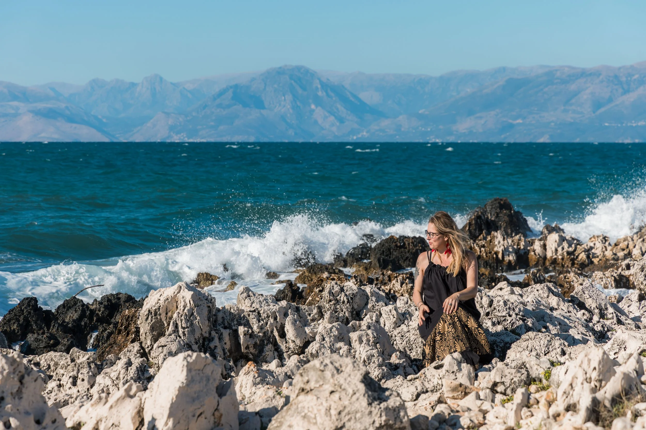 Woman sitting on rocks by the ocean with mountains in the background.