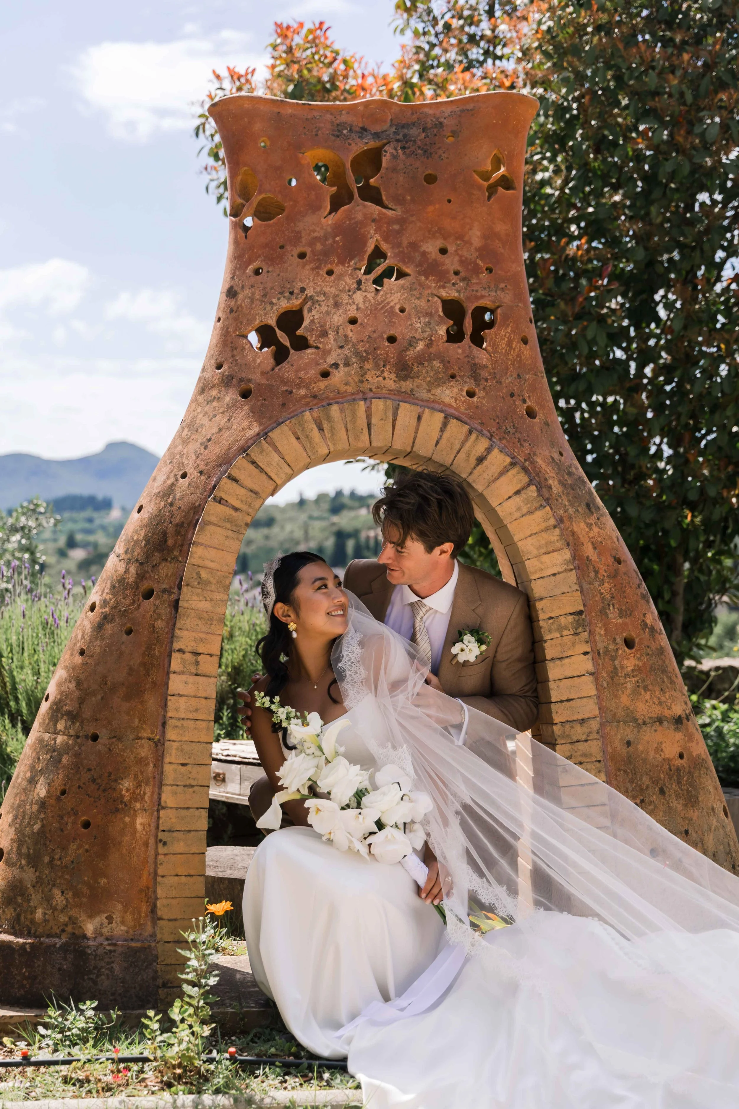 A bride and groom sitting beneath a large, rustic, outdoor sculpture with a brick arch and large, weathered clay or terra cotta structure, in a garden or countryside setting with mountains in the background.