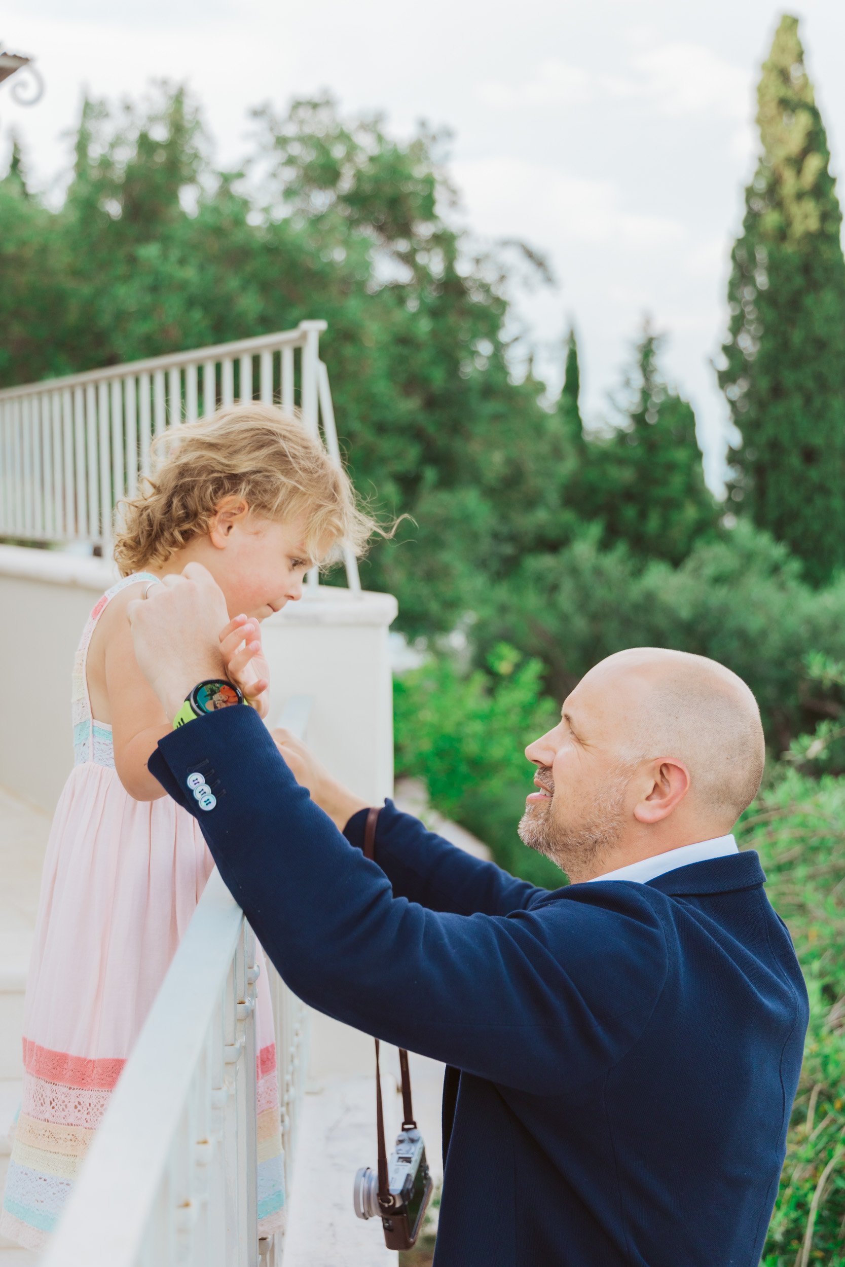 A man with a shaved head and beard smiling while holding a young girl with curly blonde hair over a white railing. The girl is wearing a pink dress with colorful stripes and is looking at the man. The background shows green trees and a partly cloudy 