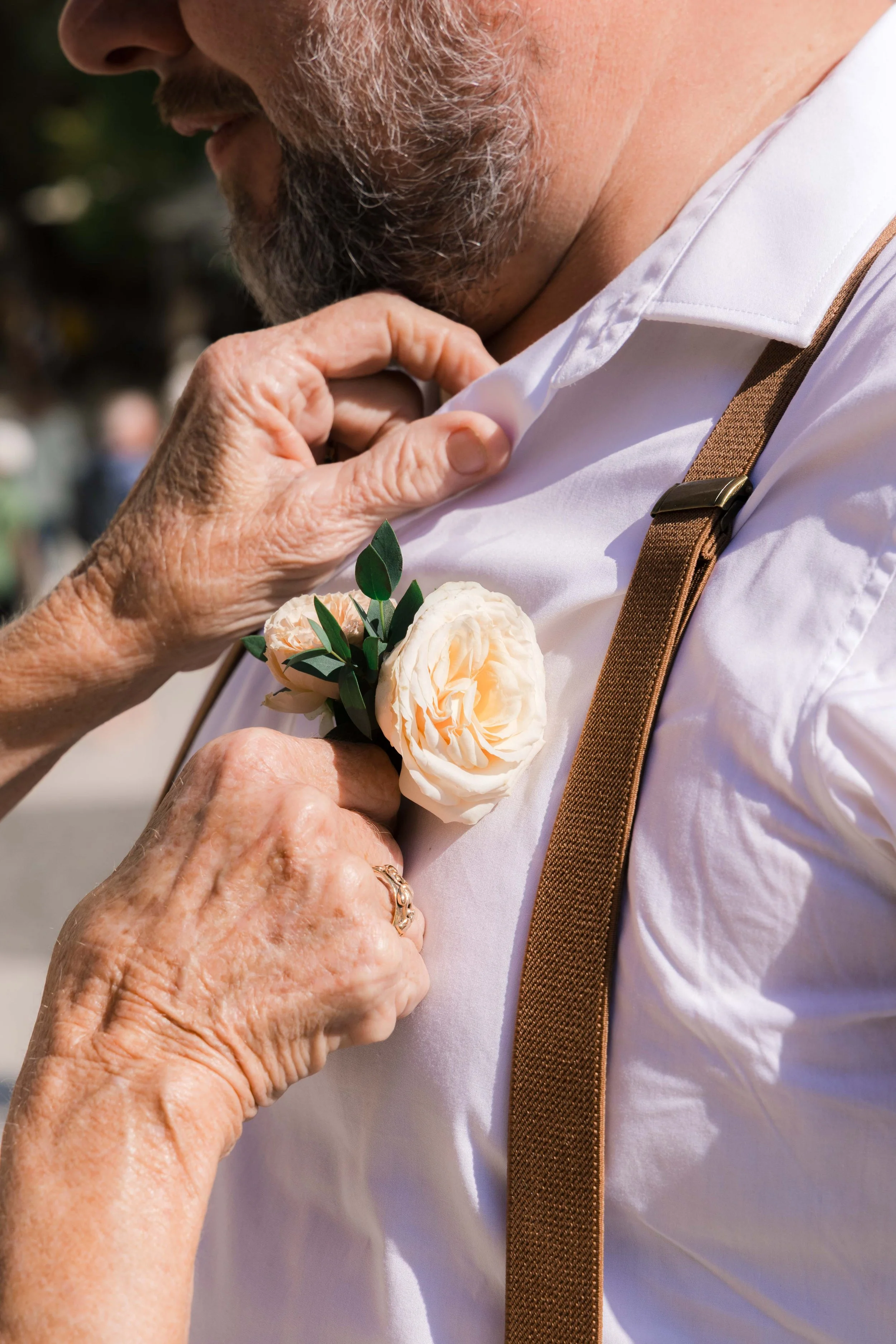 Close-up of a woman pinning a cream-colored flower boutonniere onto a man's white dress shirt, with a brown suspenders strap visible.