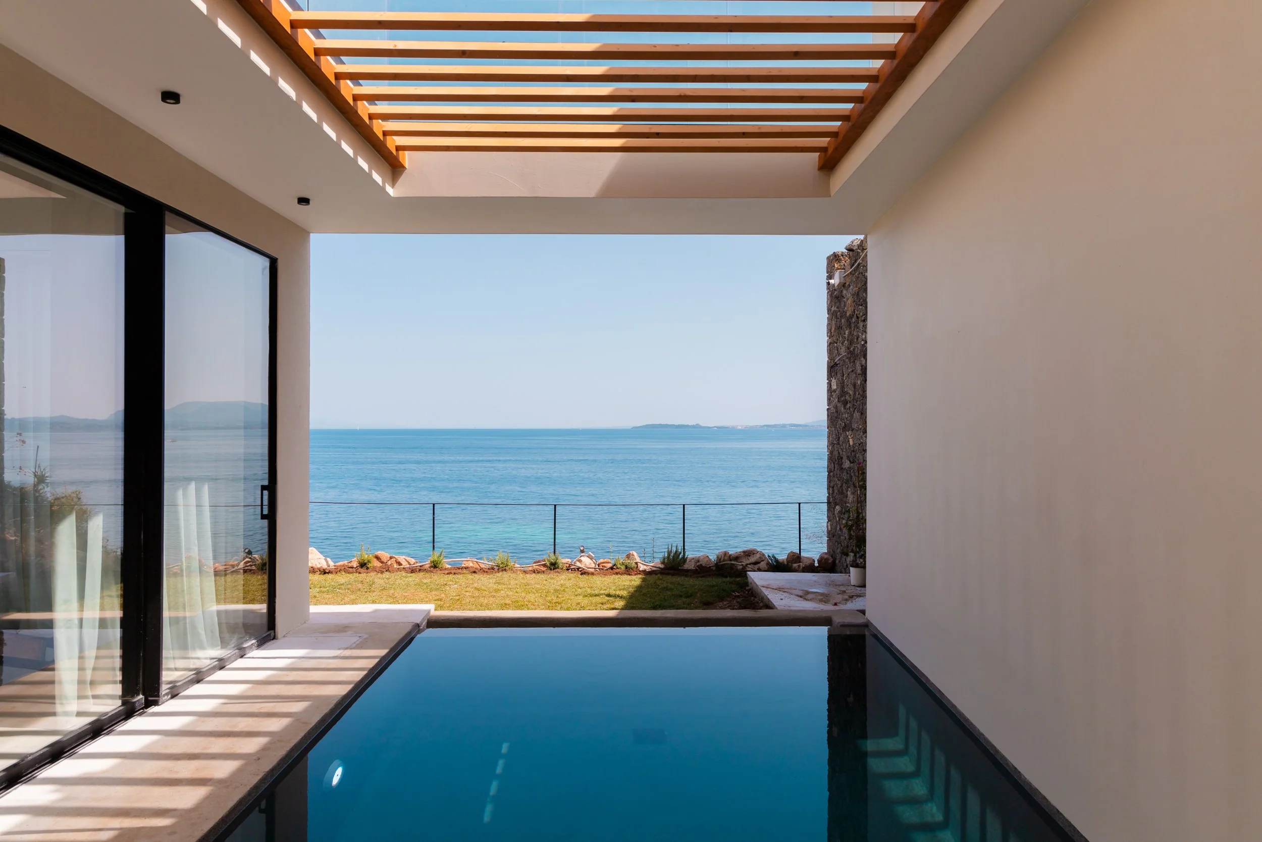 Modern indoor pool with large sliding glass doors opening to a view of the ocean and a rocky shoreline.