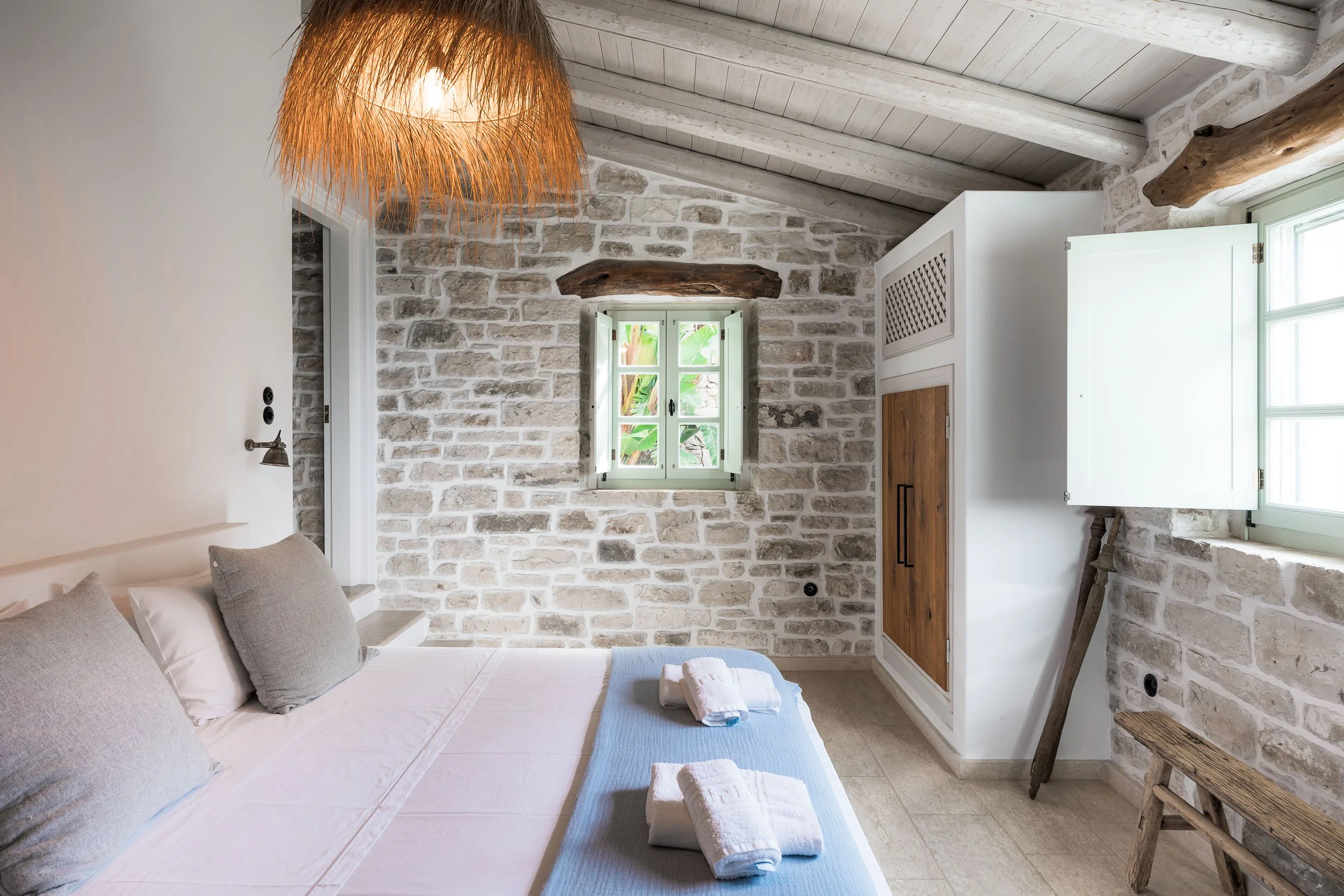 A bedroom with a stone wall, small window with shutters, and a white ceiling with exposed wooden beams. The bed has light-colored linens, pillows, and folded towels. A rustic wooden bench and a decorative hanging light fixture are also visible.