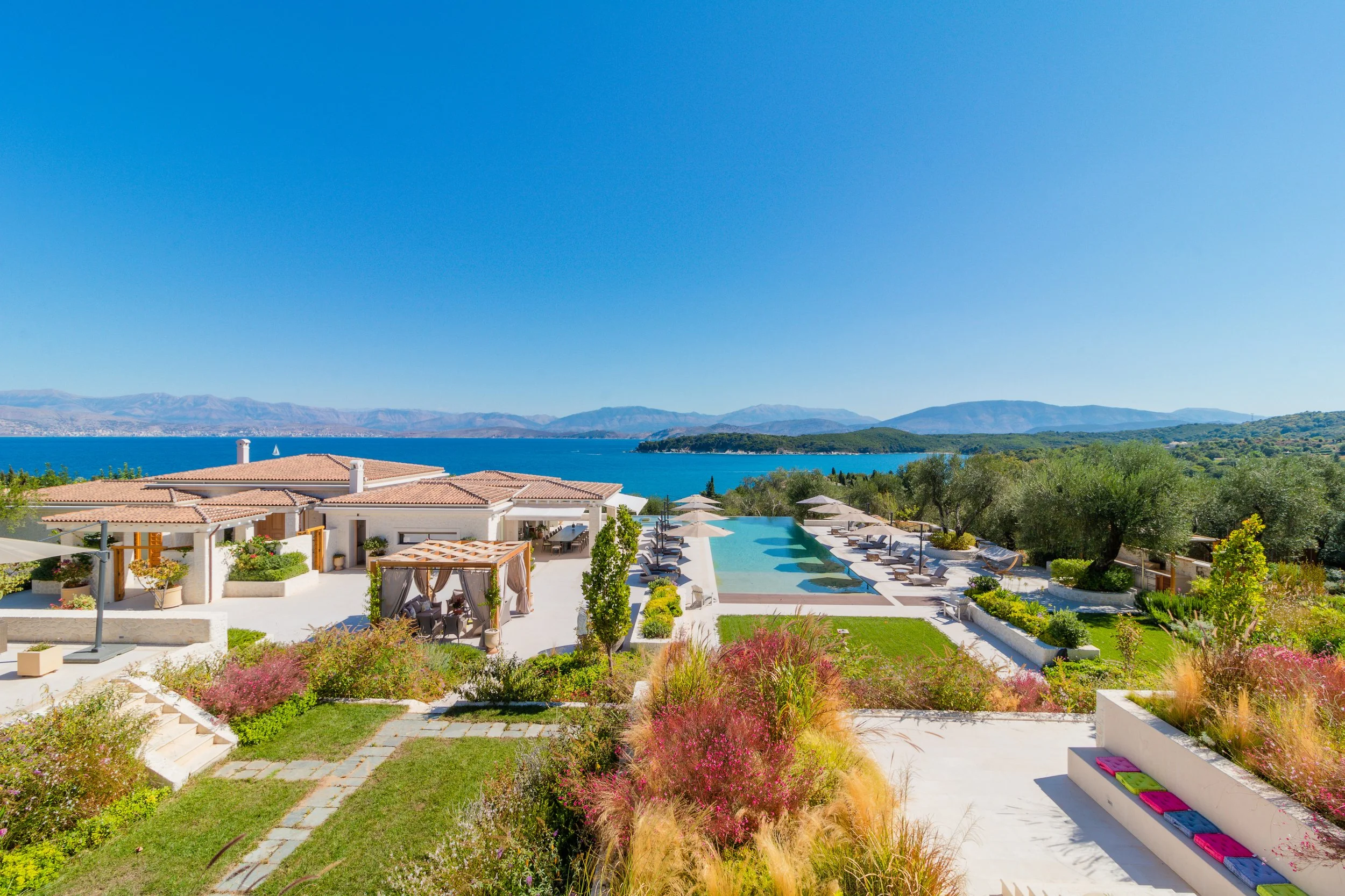 Luxury resort with a pool overlooking a lake, surrounded by greenery and mountains, under a clear blue sky.