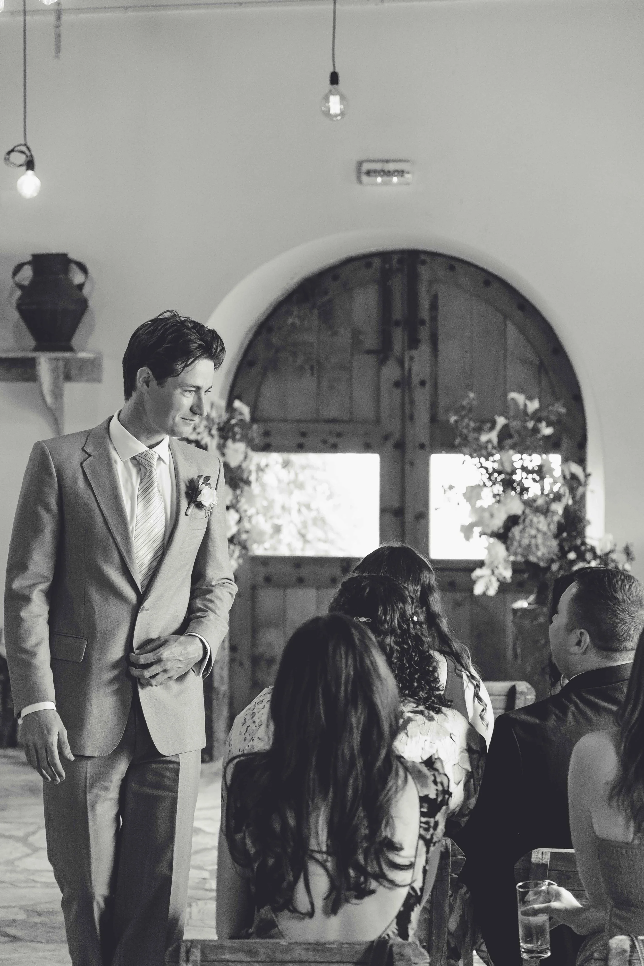 A black and white photo of a wedding reception inside a rustic venue. A groom in a suit with a boutonniere is smiling and facing a seated group of women and men. The background features large wooden doors, floral arrangements, and hanging light bulbs