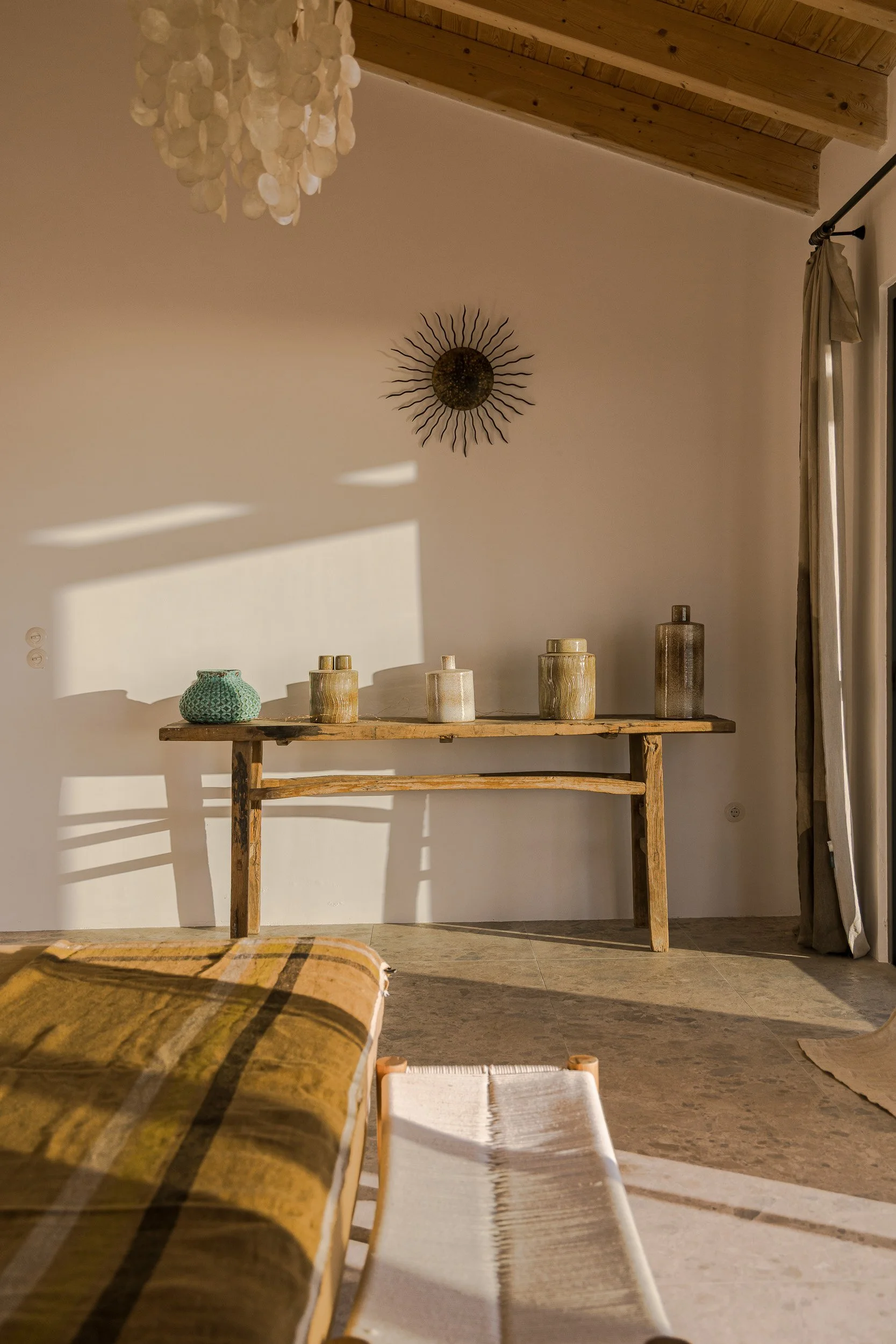 Sunlight casts shadows on a white wall with a sun sculpture. A wooden table holds five decorative vases. Part of a bed with a plaid blanket is visible in the foreground, along with a woven bench and curtains beside a sliding glass door.