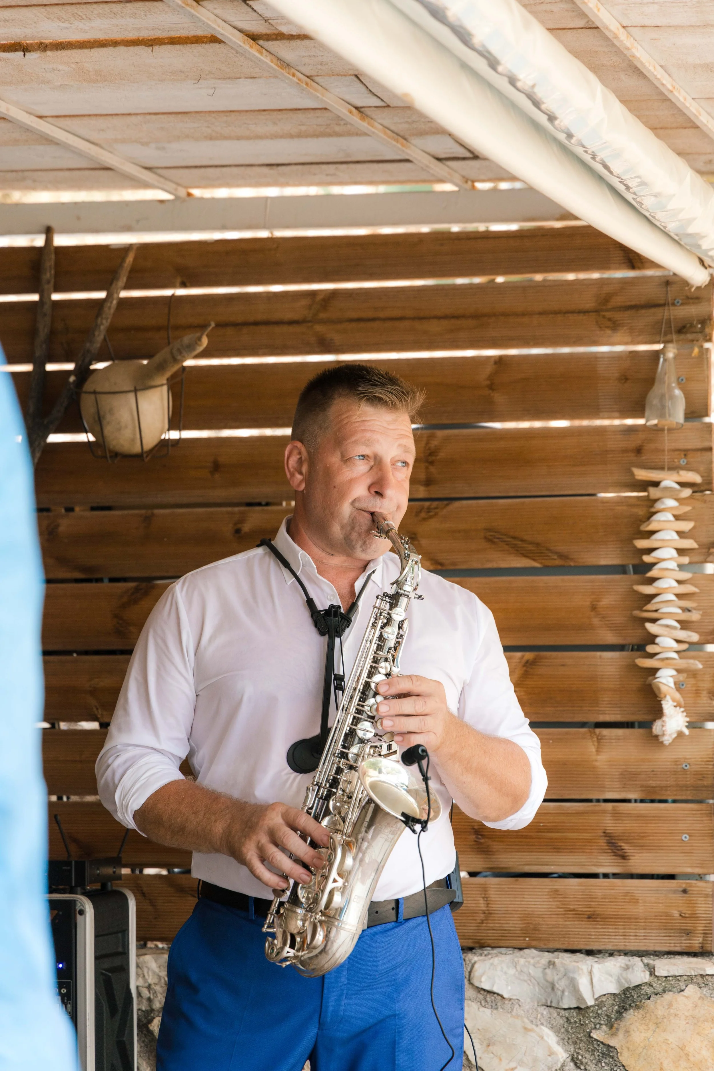A man in a white shirt playing a saxophone in an outdoor setting with wooden walls and decorative shells.