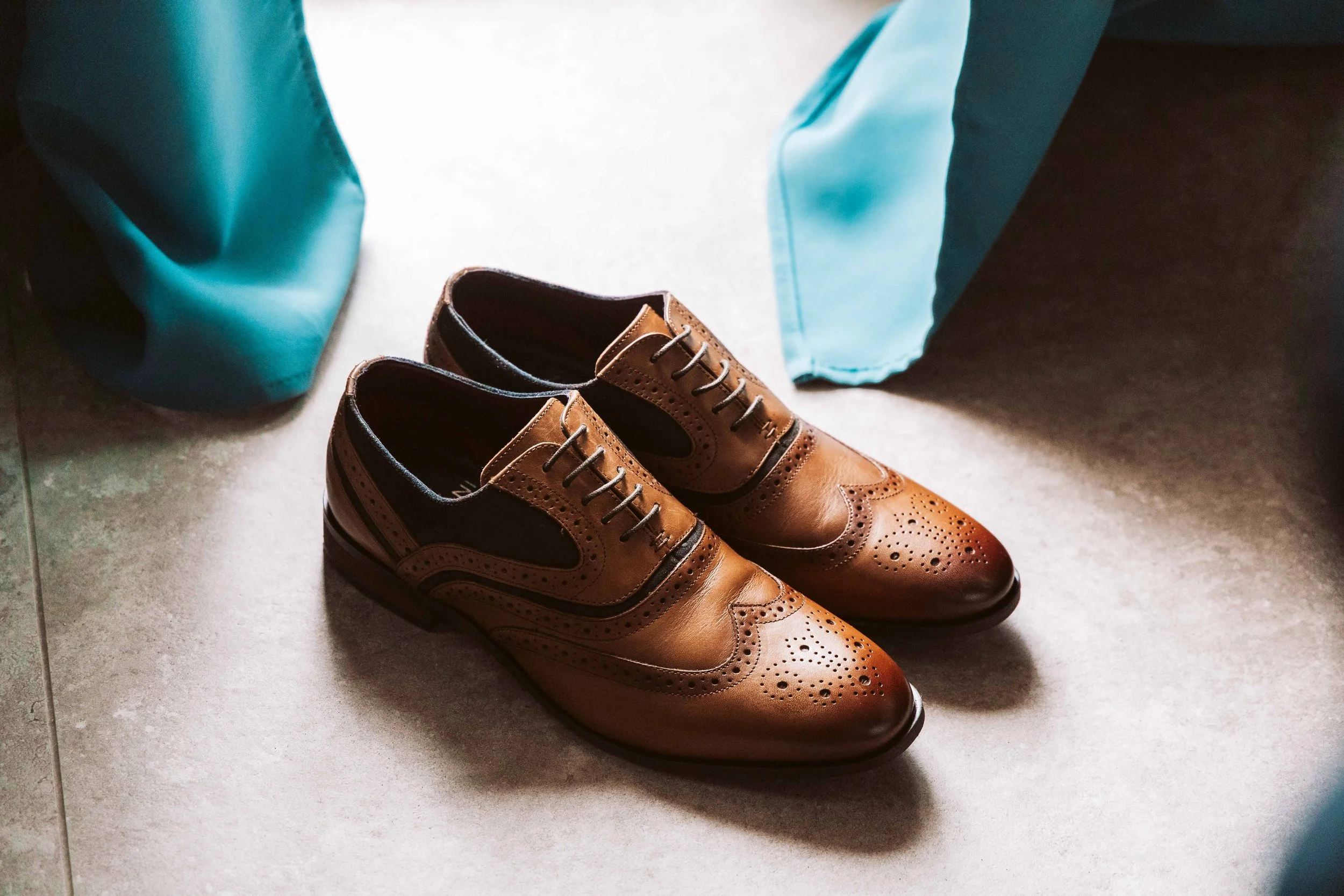 Pair of brown and black leather brogue dress shoes on a light-colored floor, partially under a turquoise cloth or curtain.