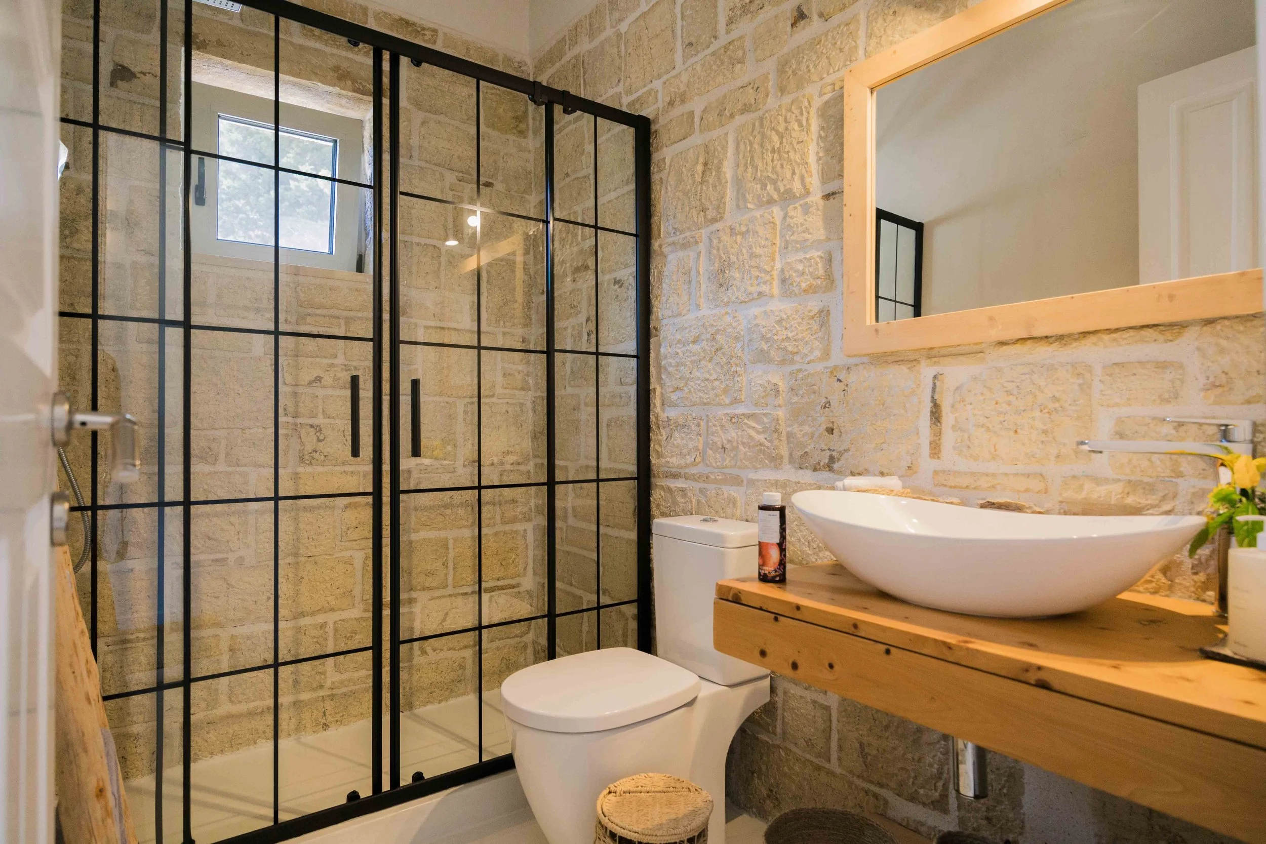 Bathroom with stone walls, glass shower enclosure, white toilet, and a wooden vanity with a white vessel sink and mirror.