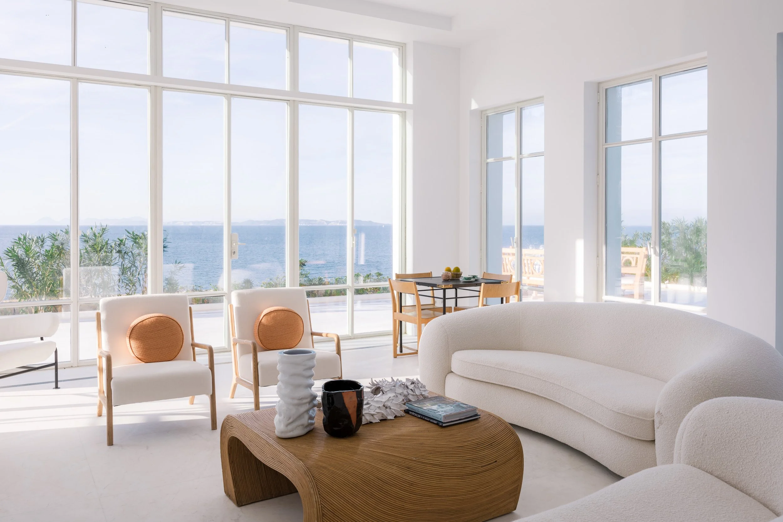 Bright living room with large windows showing ocean view, white sofa, two white chairs with orange cushions, wooden coffee table with decorative vases, and small dining area with table and chairs near the windows.