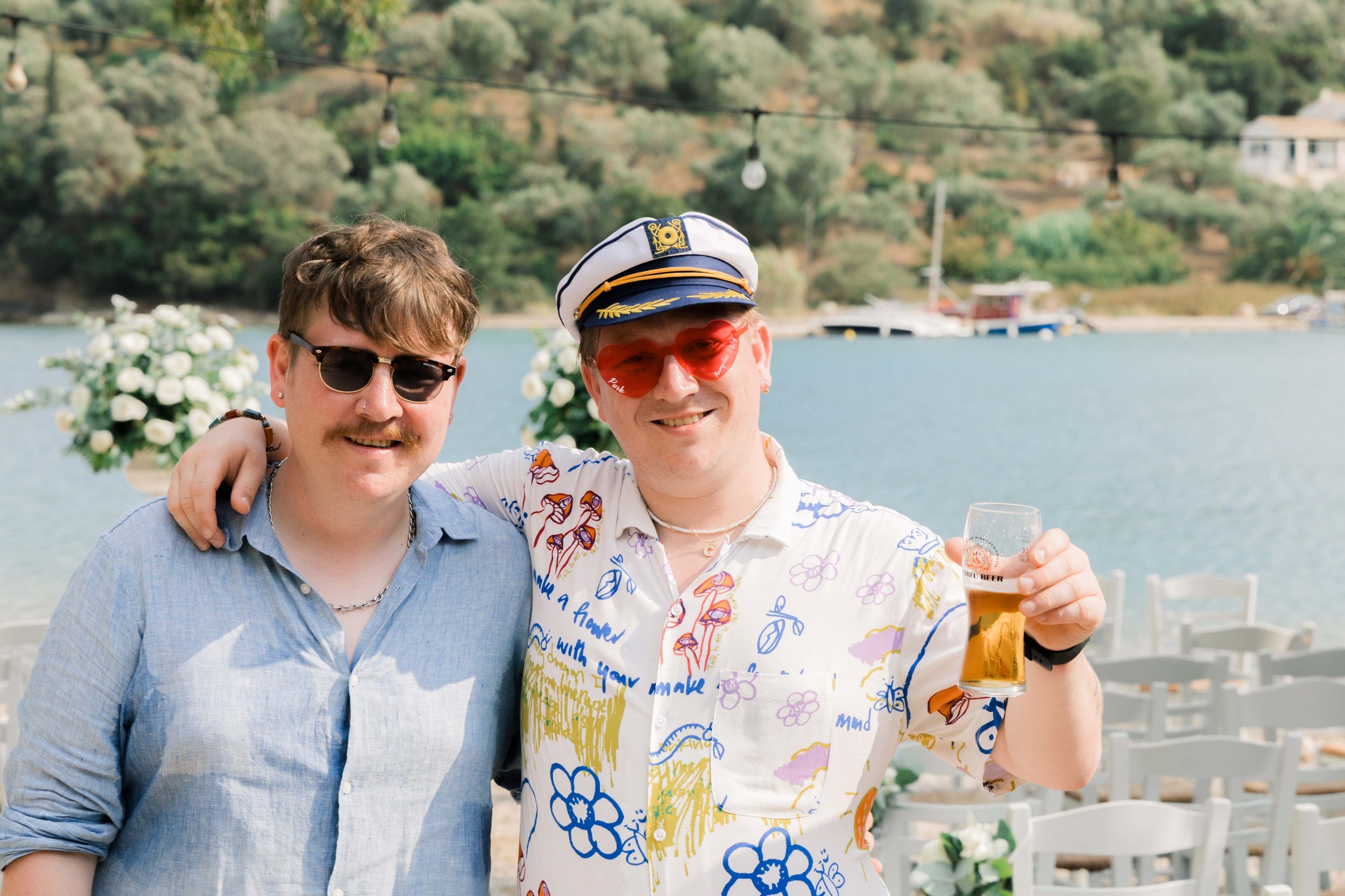 Two men smiling with arms around each other at an outdoor lakeside event, one holding a glass of beer, with boats and hills in the background, decorated with flowers and string lights.