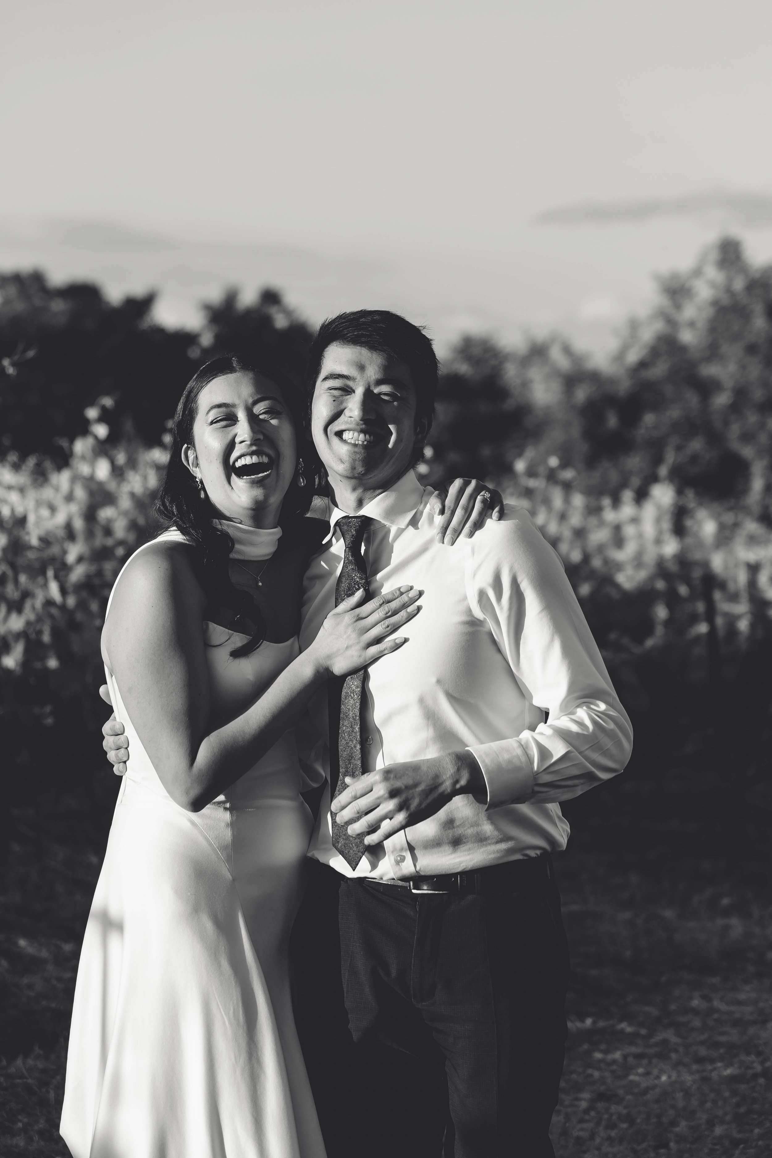 Black and white photo of a happy couple outdoors, embracing and smiling, with trees in the background.