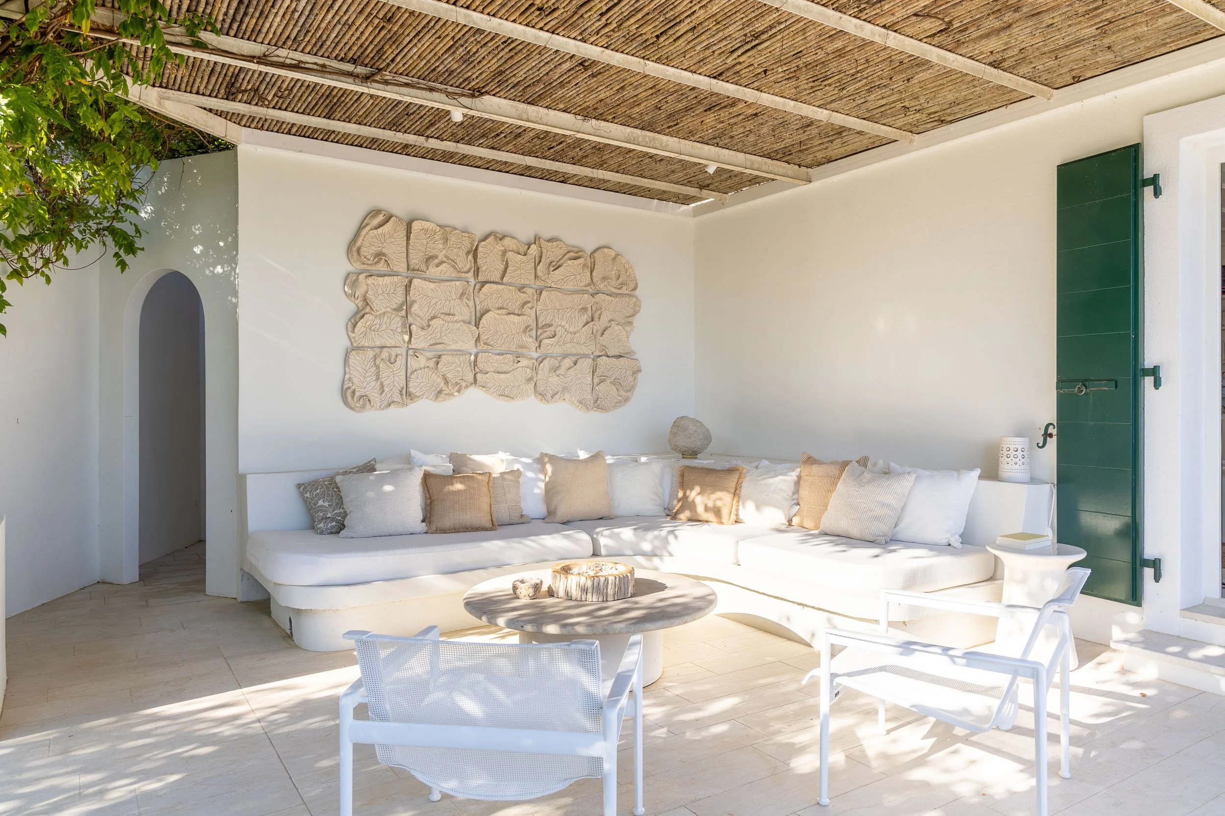 Outdoor seating area with white L-shaped sofa, wooden table, and white chairs, decorated with neutral-colored pillows, against a white wall with beige wall art, under a thatched roof.
