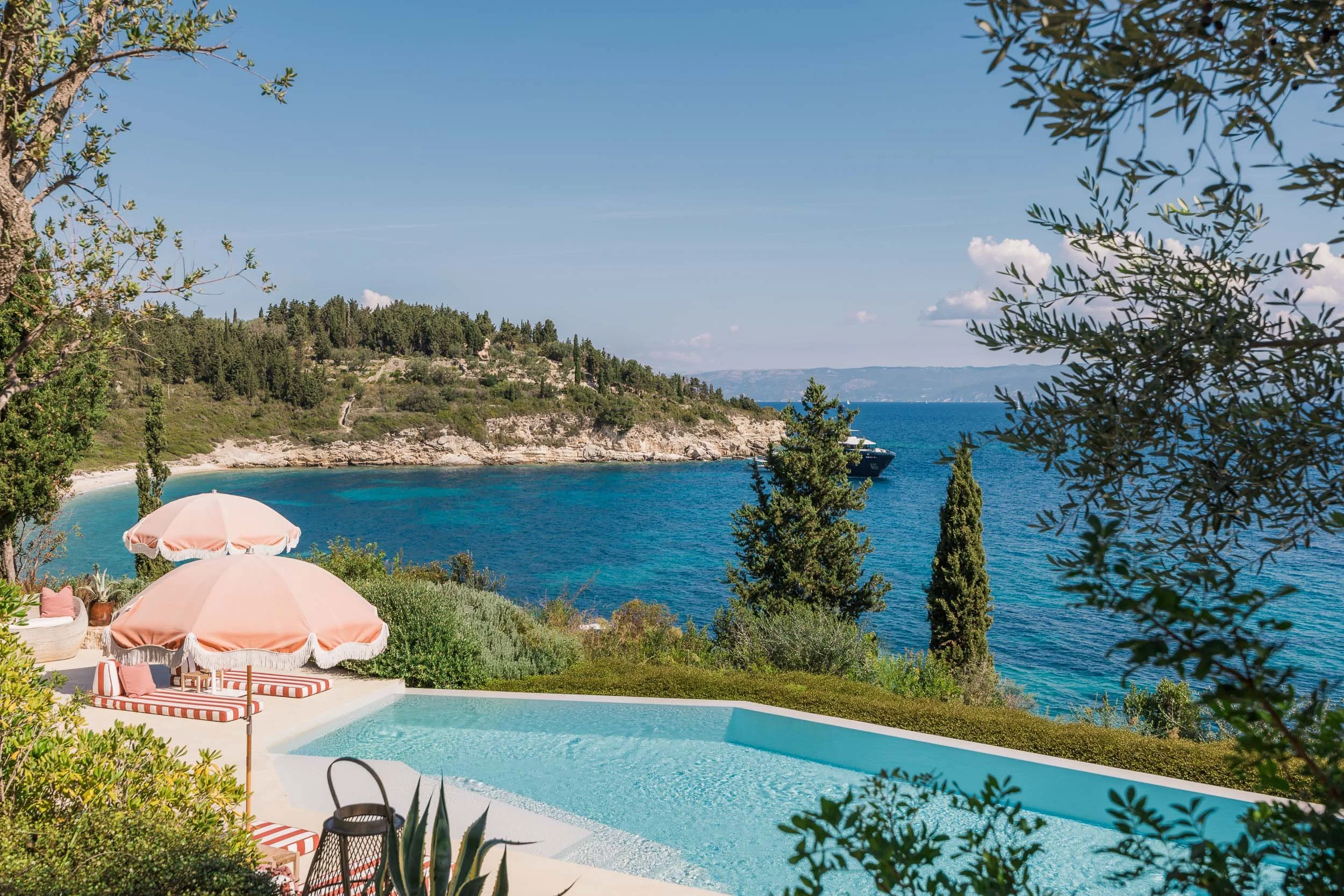 A swimming pool overlooking a bay with a boat, surrounded by trees and greenery, with pink umbrellas and lounge chairs nearby.