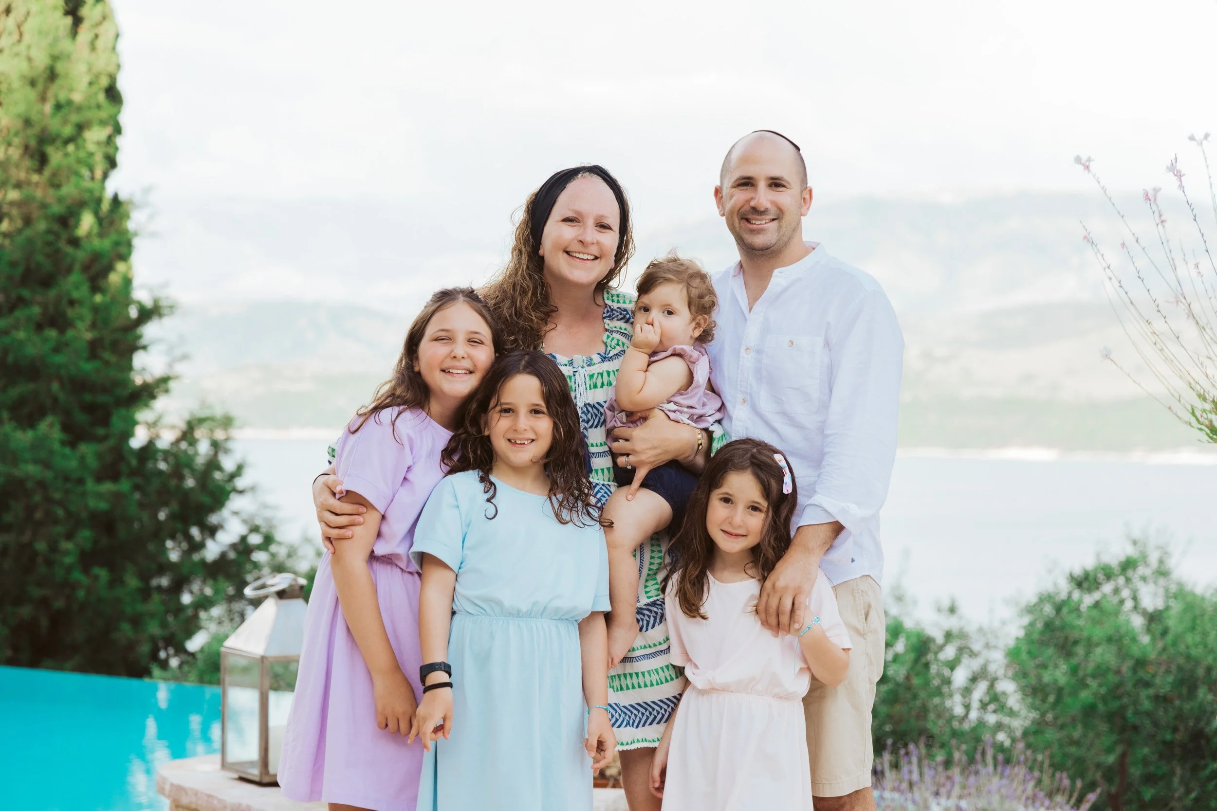 A family of seven posing outdoors near a body of water, with mountains in the background. The family includes two adults and five children, all smiling at the camera.