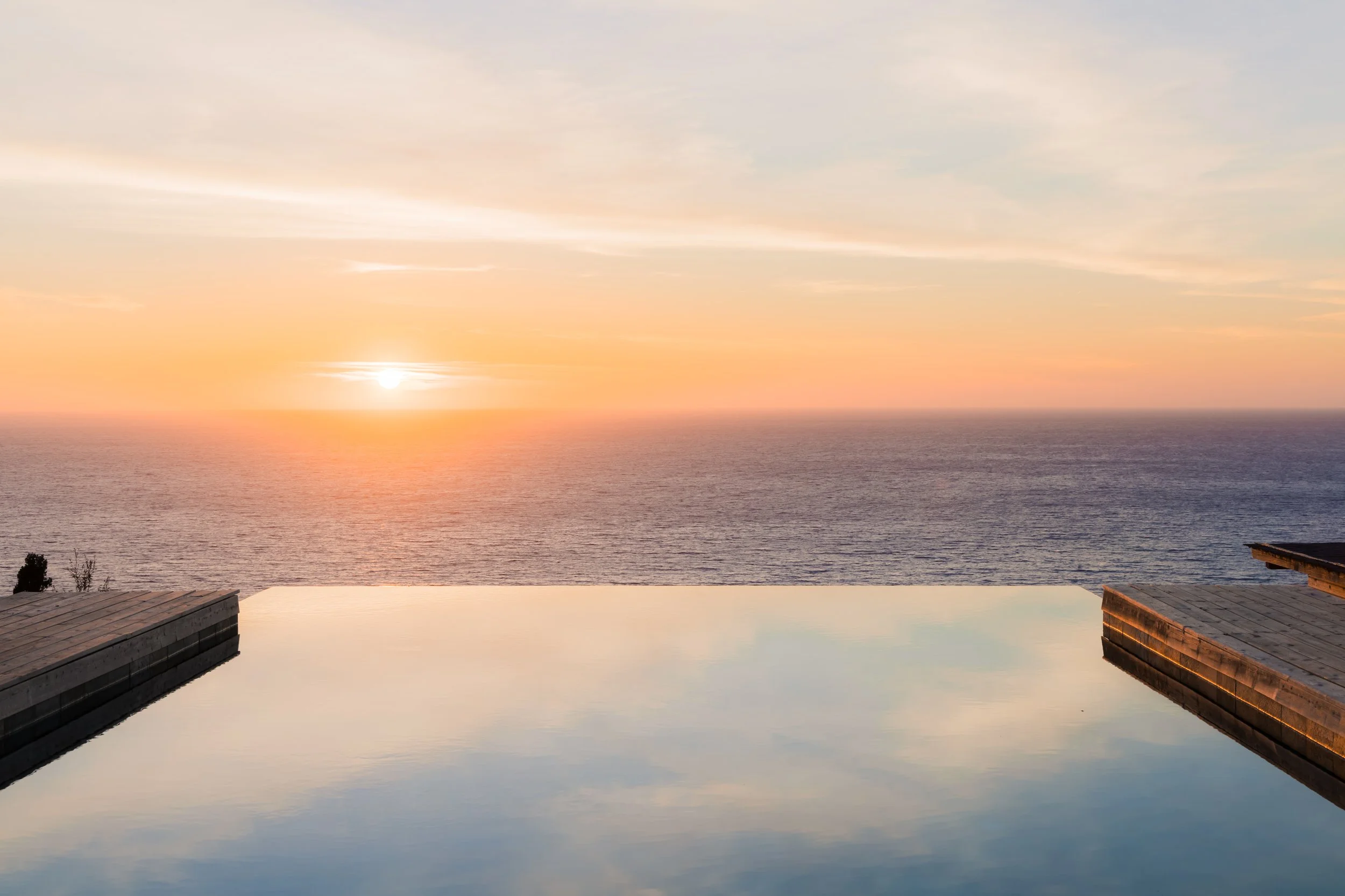Sunset view over the ocean with an infinity pool in the foreground, reflecting the sky. Wooden deck edges on either side of the pool.