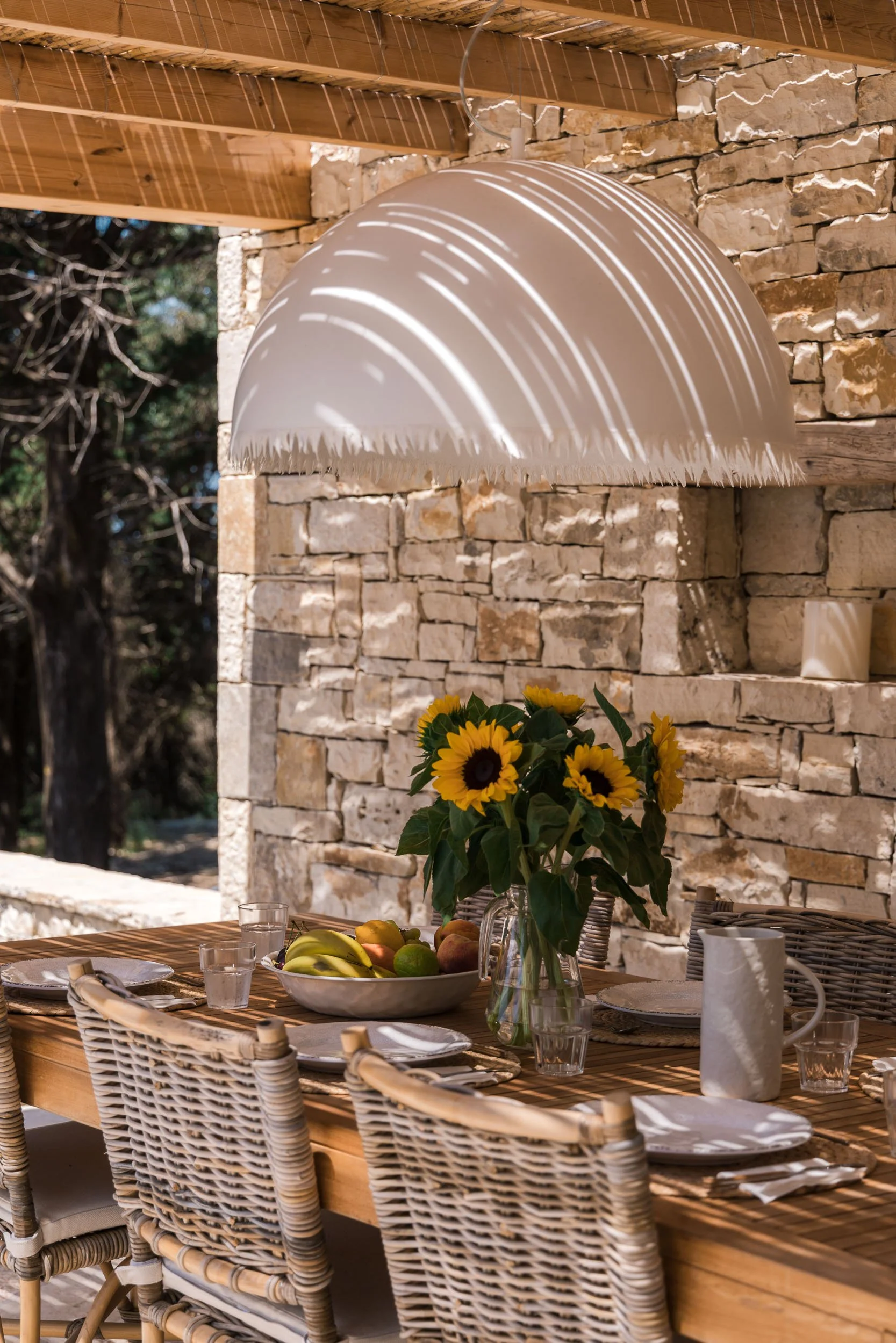 A wooden outdoor dining table with plates, glasses, and a pitcher. A vase with sunflower flowers and a bowl of fruit, including bananas, pears, and apples. Wicker chairs around the table. A large hanging pendant light casting striped shadows. Stone w