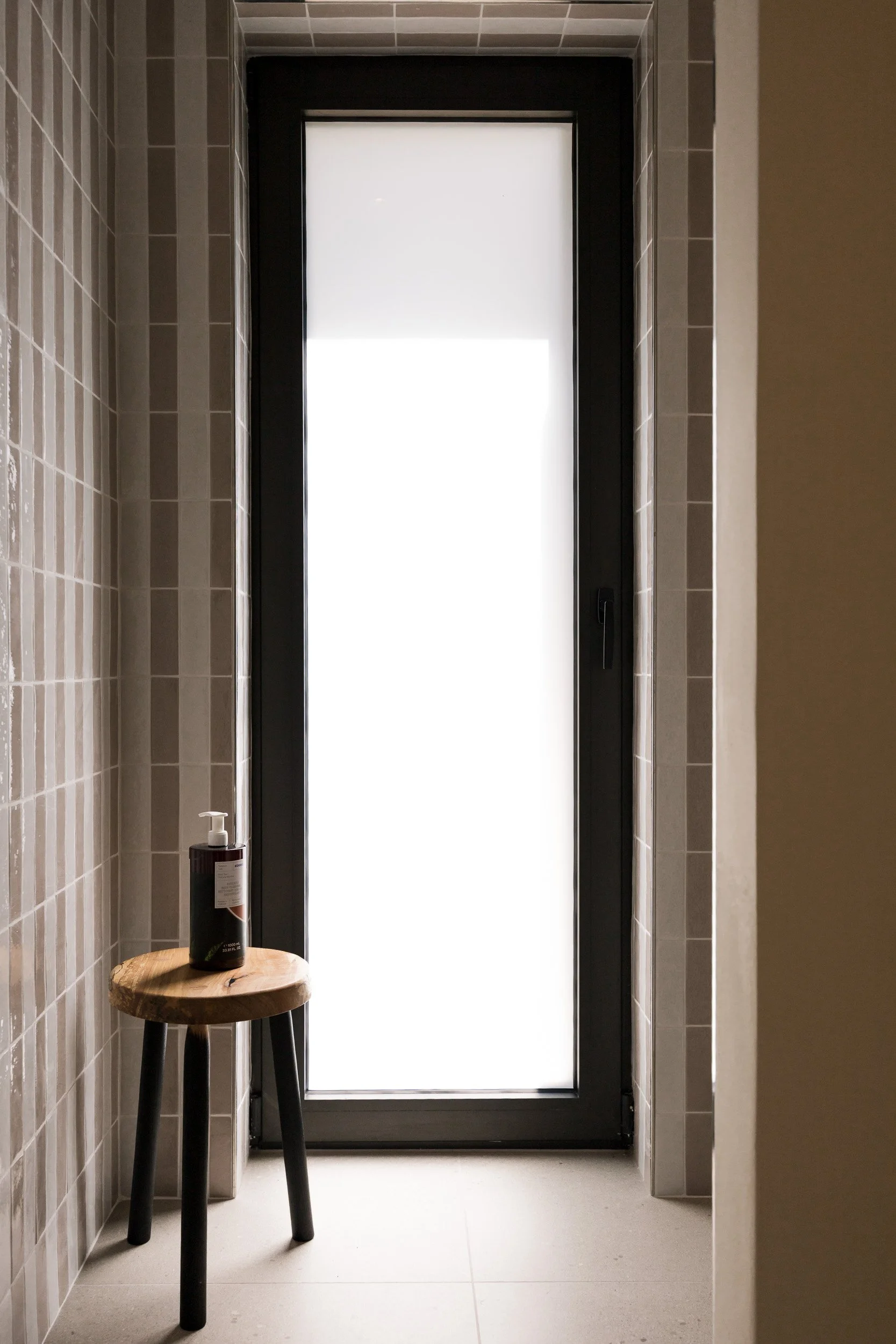 A narrow bathroom or shower room with beige tile walls, a frosted glass door, a small wooden stool with black legs, and a bottle of soap or lotion on top of the stool.