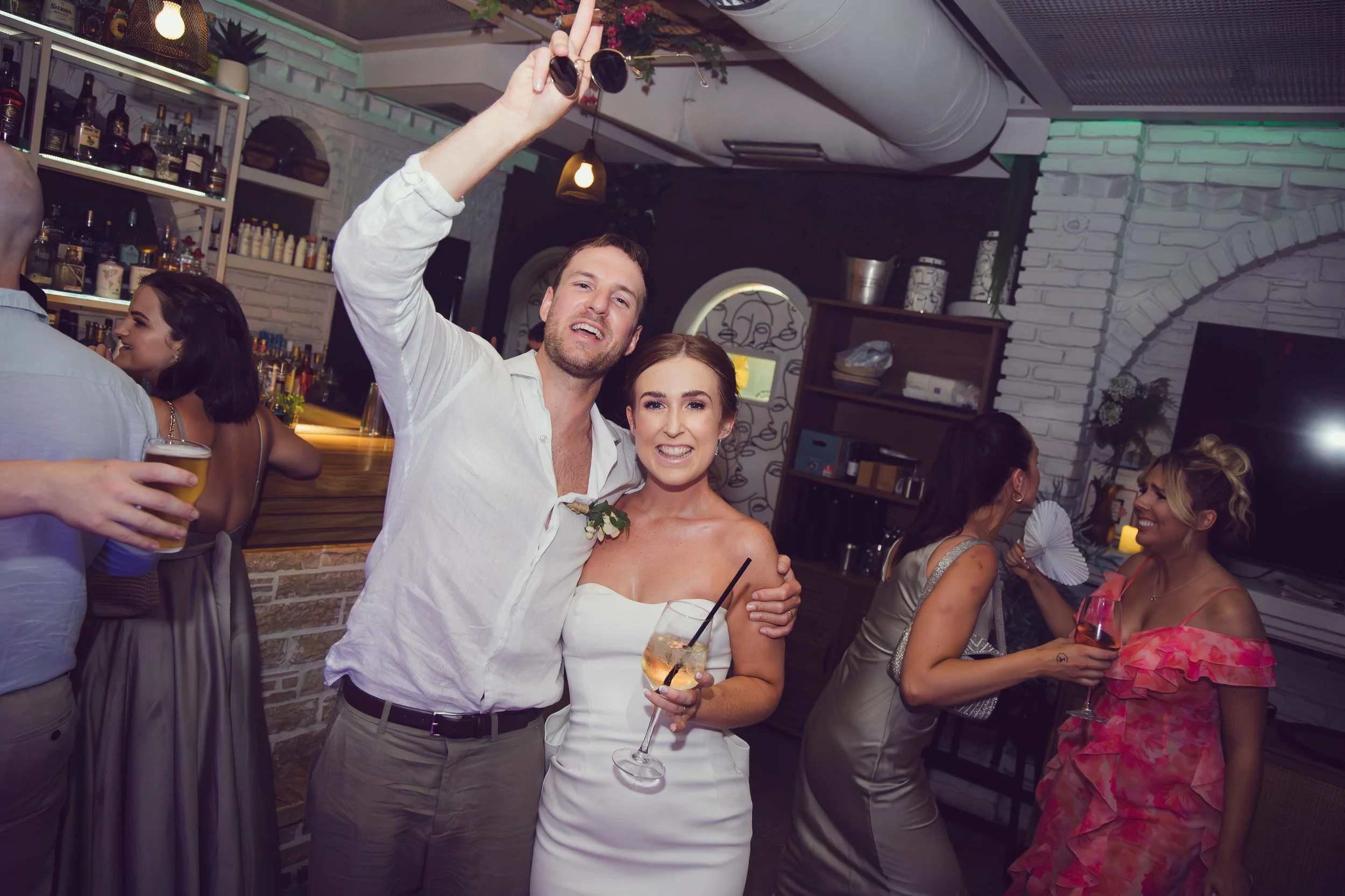 People celebrating at an indoor party, with a man making a peace sign and standing next to a woman holding a cocktail, while other guests chat and drink in the background.