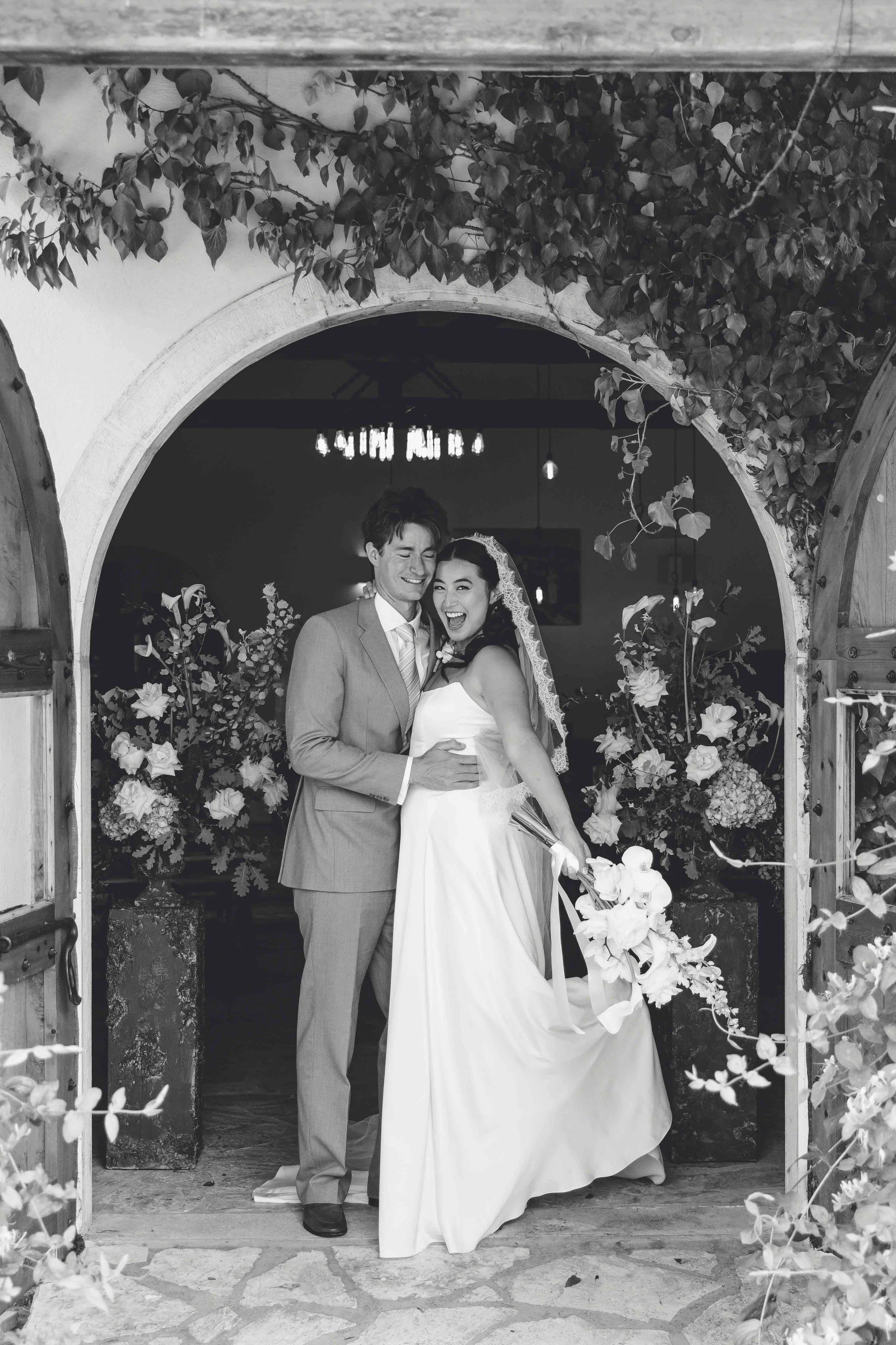 Black and white photo of a bride and groom smiling inside a decorated archway with flowers.