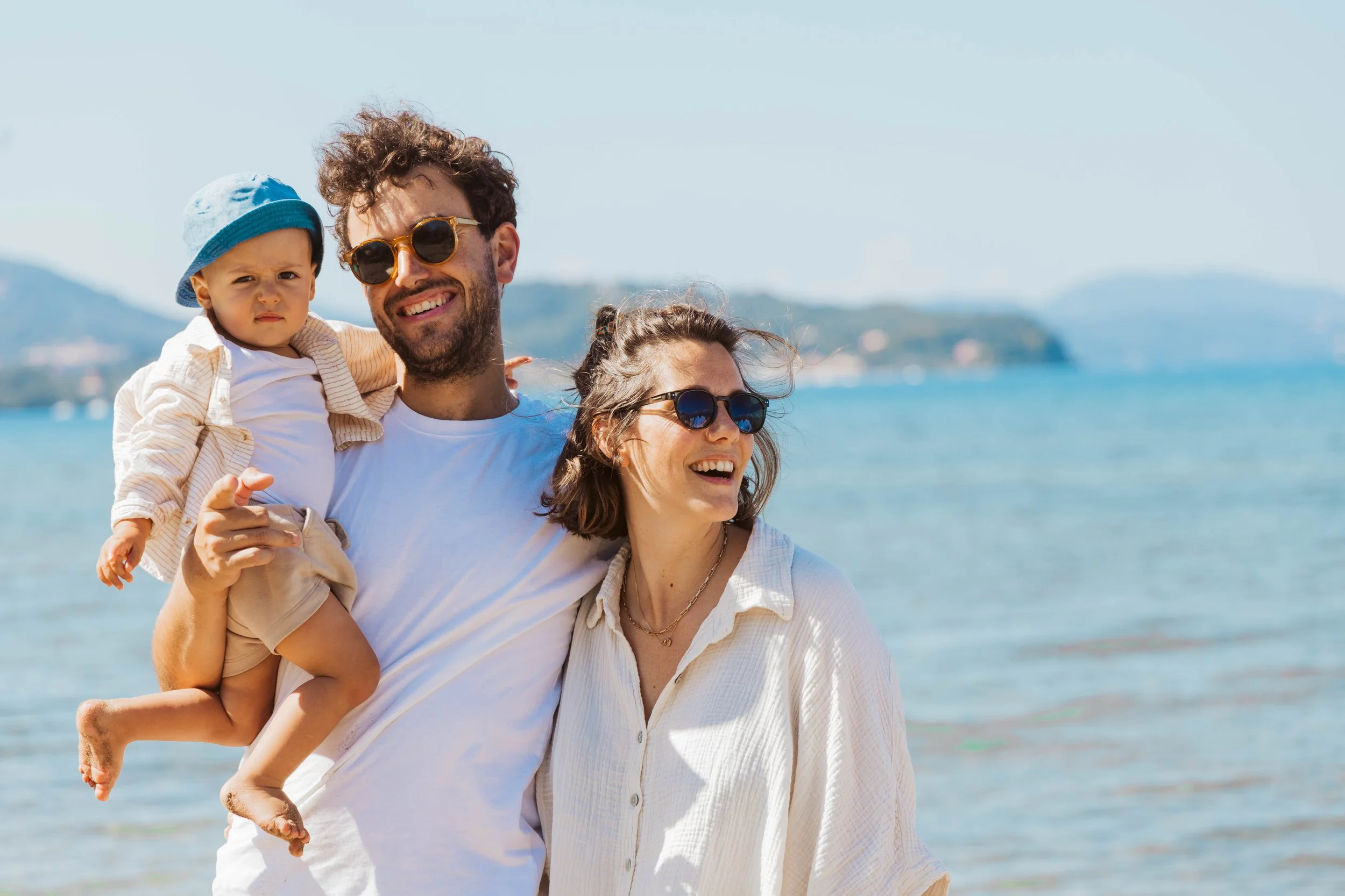 Happy family at the beach, with a man carrying a toddler and a woman smiling, all wearing sunglasses and light clothing, with water and hills in the background.