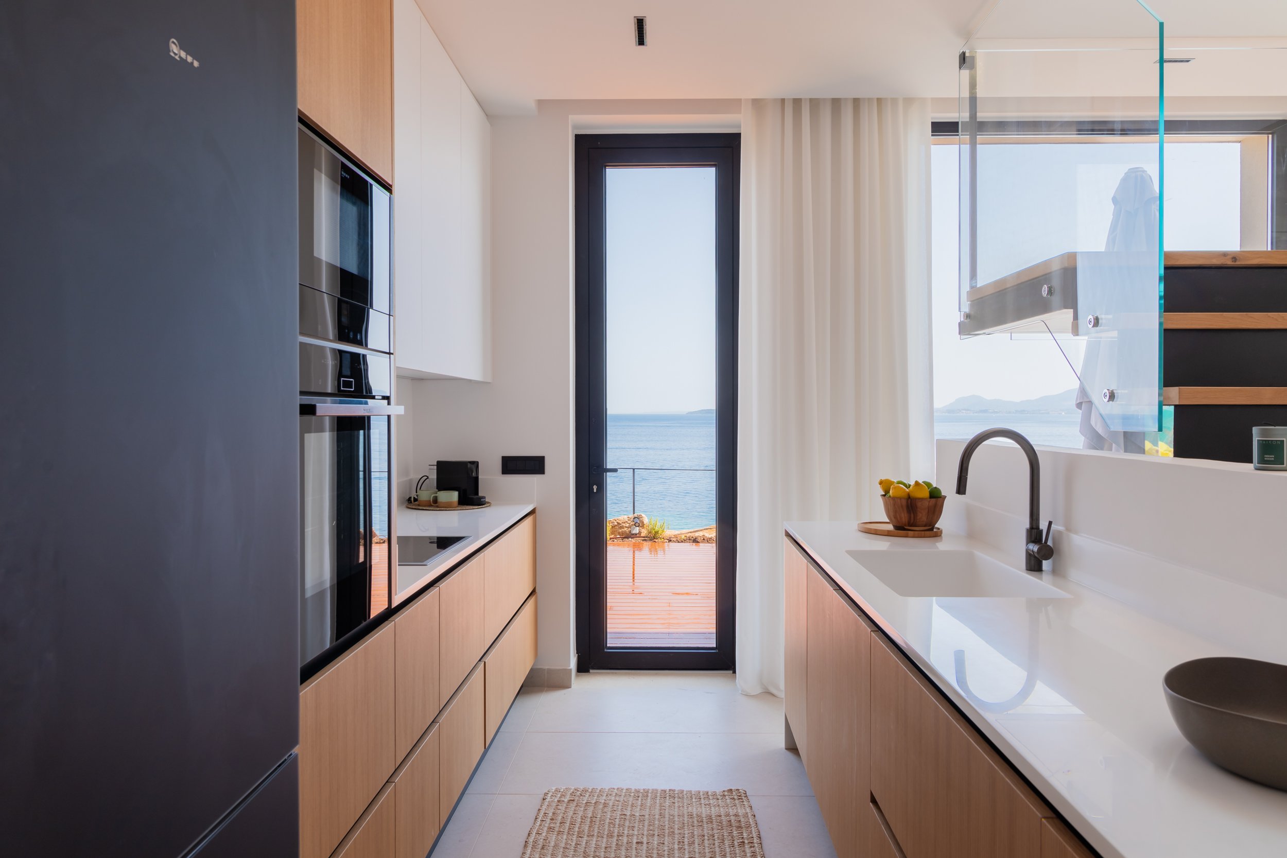 Modern kitchen with a view of the ocean through a glass door, featuring light wood cabinets, a white countertop, a sink, and minimal decor.