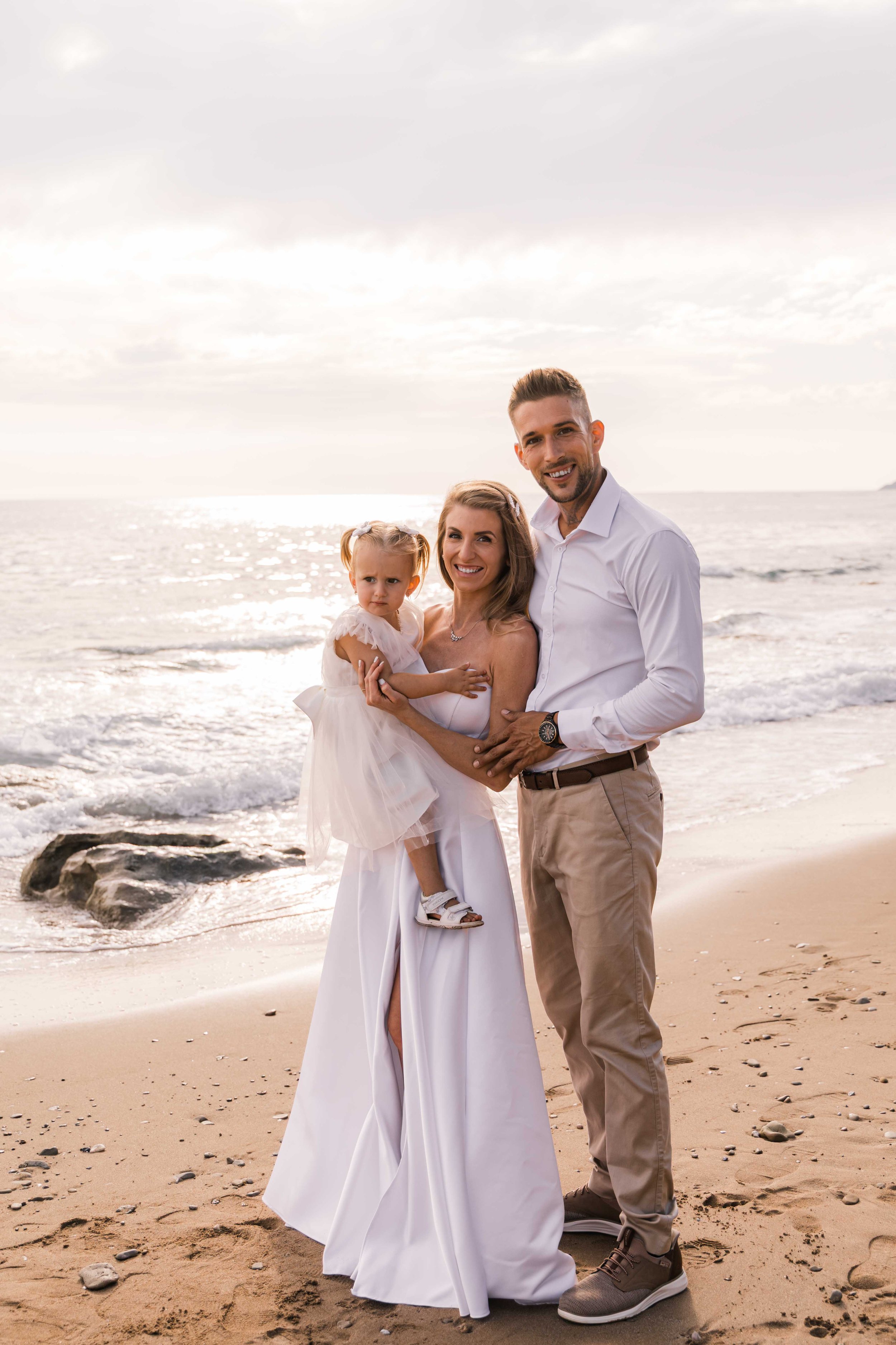 Family standing on a beach with ocean and sky in the background, sunlight reflecting on water, and rocks on the sand.