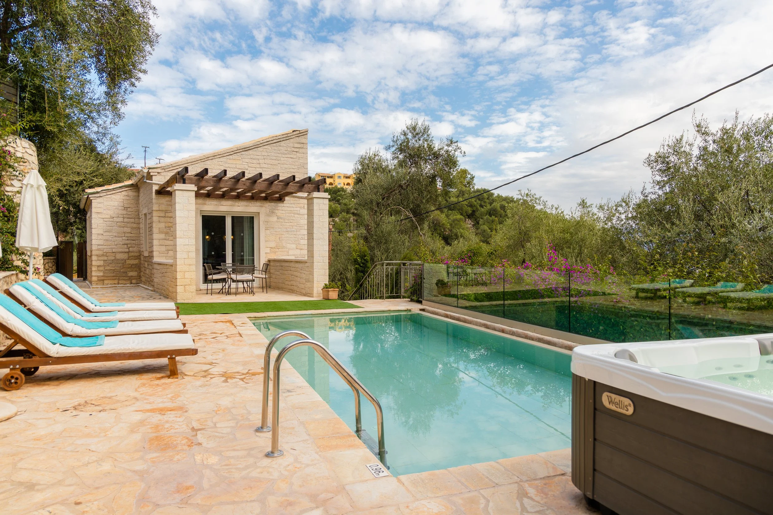 A backyard with a swimming pool, poolside lounge chairs with blue cushions, a hot tub, and a small patio area with a table and chairs, surrounded by trees and greenery under a partly cloudy sky.