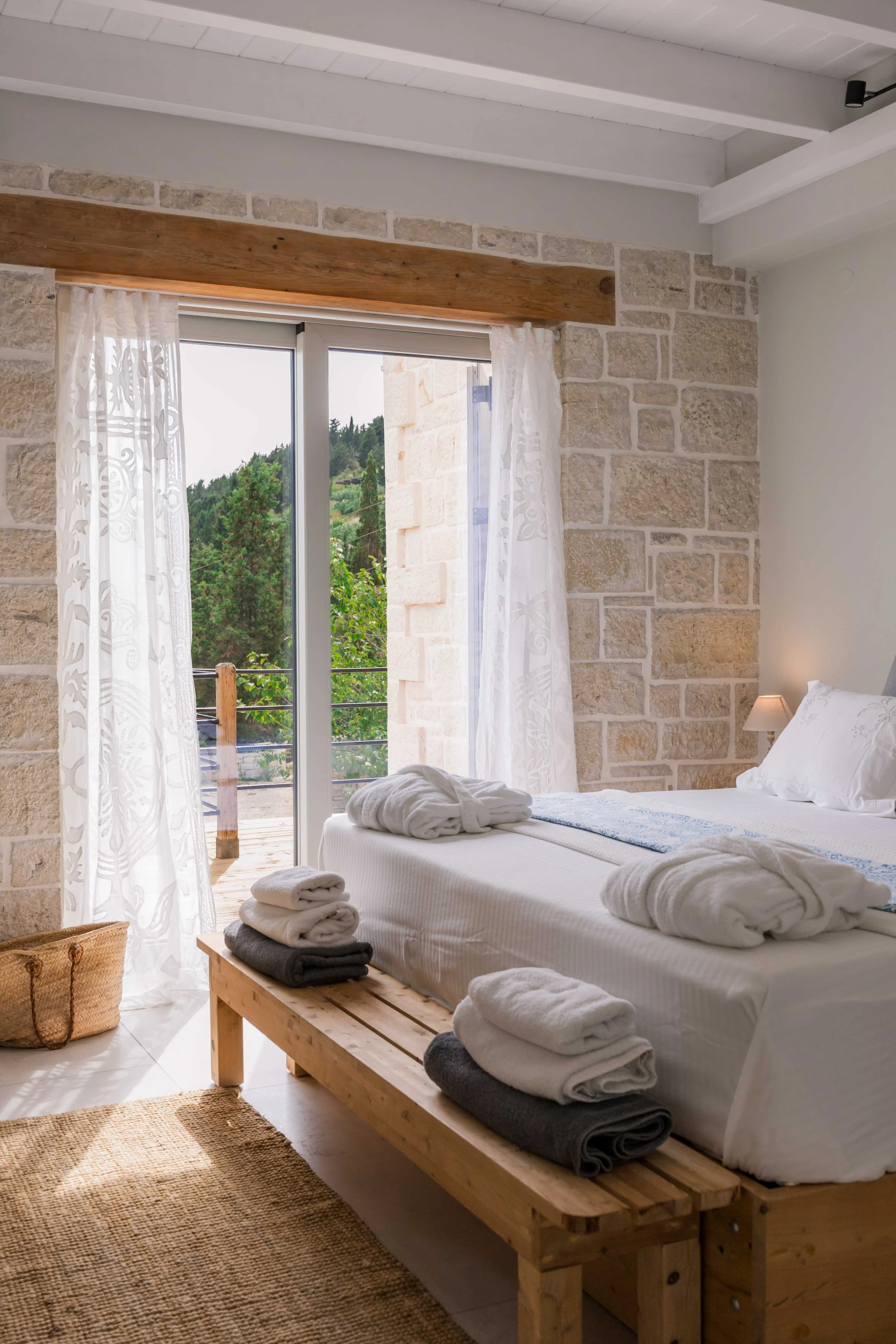 A cozy bedroom with a stone accent wall, a large window with white curtains, a wooden bed with white linens and towels, and a balcony view of trees.