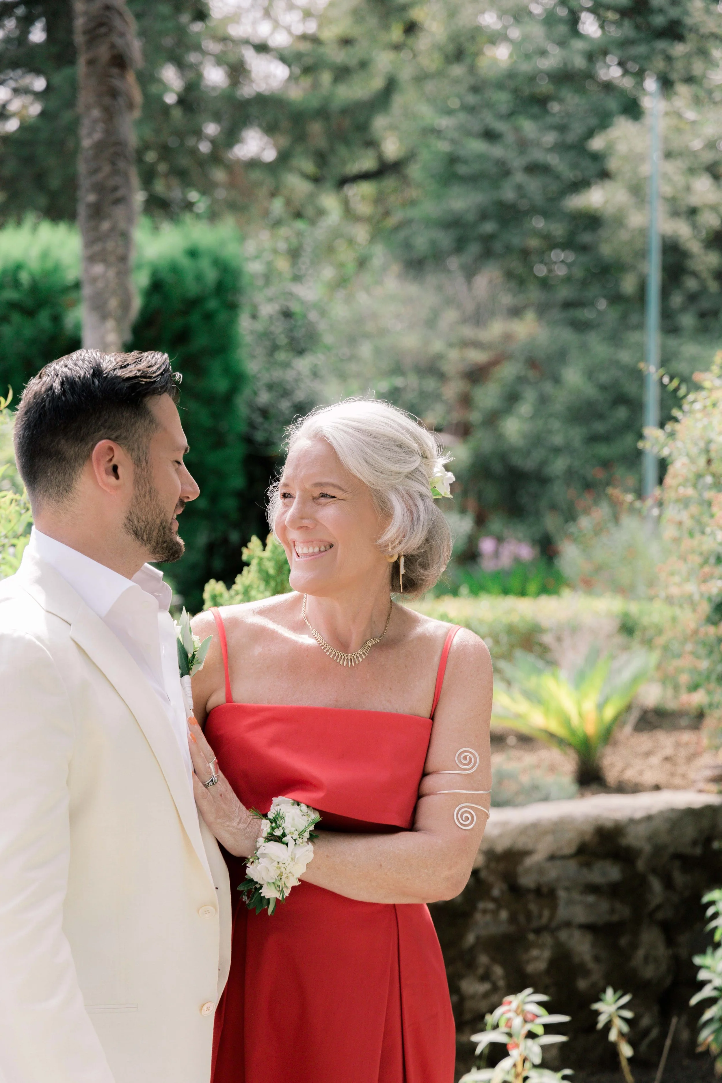 A bride in a red dress smiling and looking at a groom in a white suit, outdoors in a garden setting.