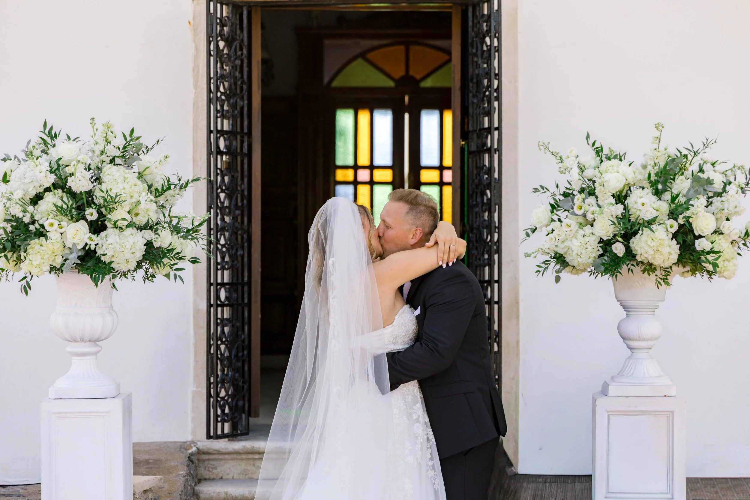 Bride and groom embracing and kissing at their wedding, standing in front of a church entrance with stained glass windows, flanked by large floral arrangements in white vases.