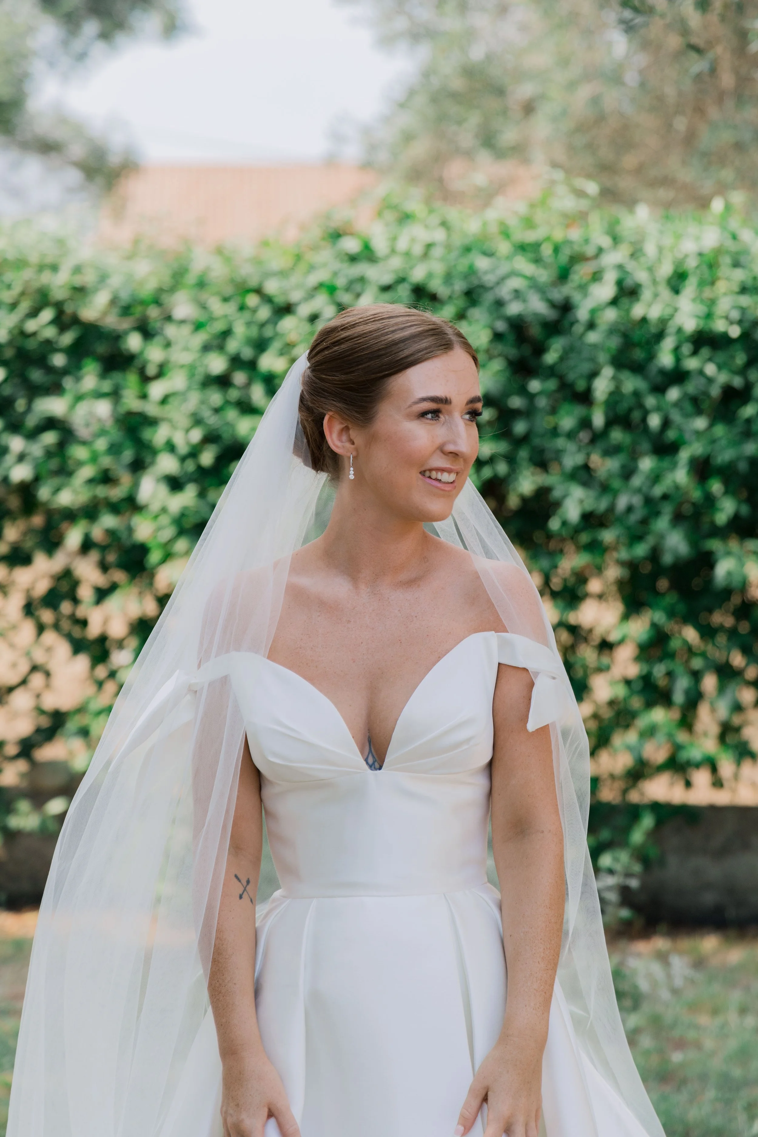 A bride in a white wedding dress and veil smiling outdoors with green shrubbery in the background.