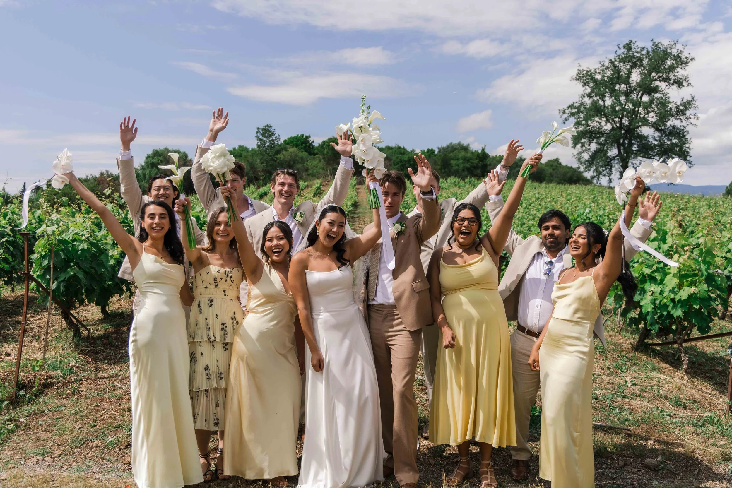 A group of people celebrating outdoors on a sunny day, holding bouquets of white flowers, with green vineyard rows and a large tree in the background.