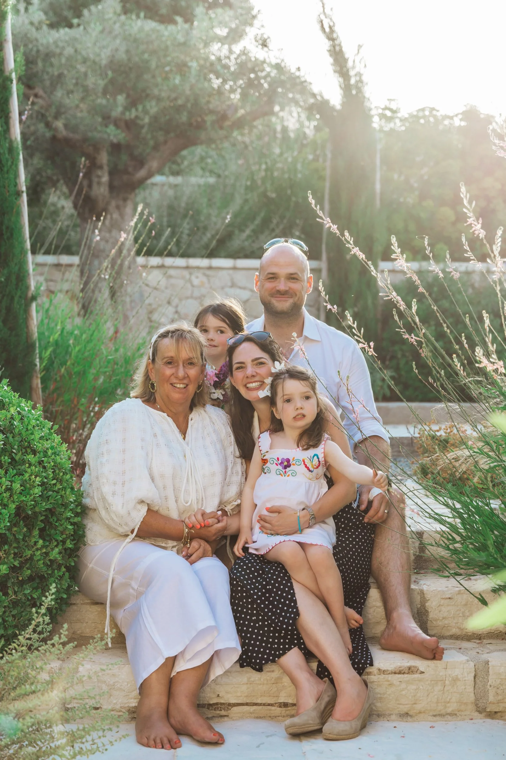 A family of five sitting outdoors on stone steps surrounded by greenery during sunset. The family includes an elderly woman, a young woman, a man, and two young girls. The family is smiling and enjoying the moment together.