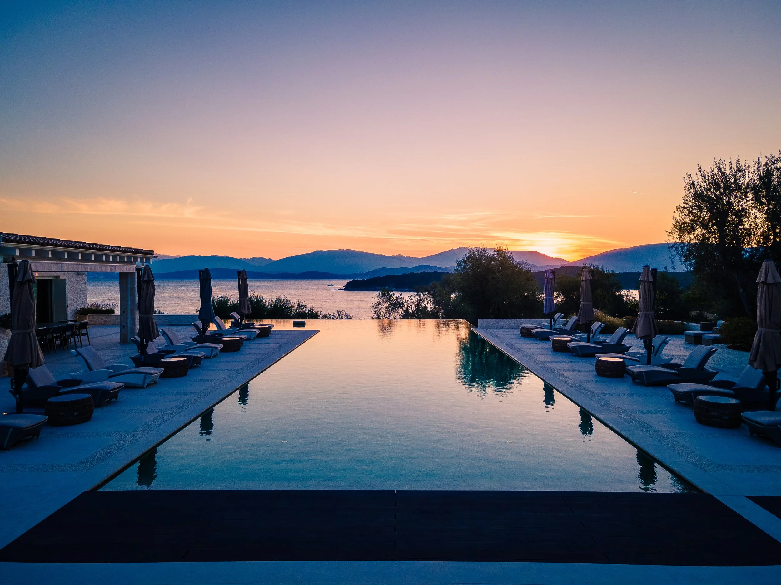 Sunset view over a swimming pool with lounge chairs and umbrellas, overlooking a lake and mountains in the distance.