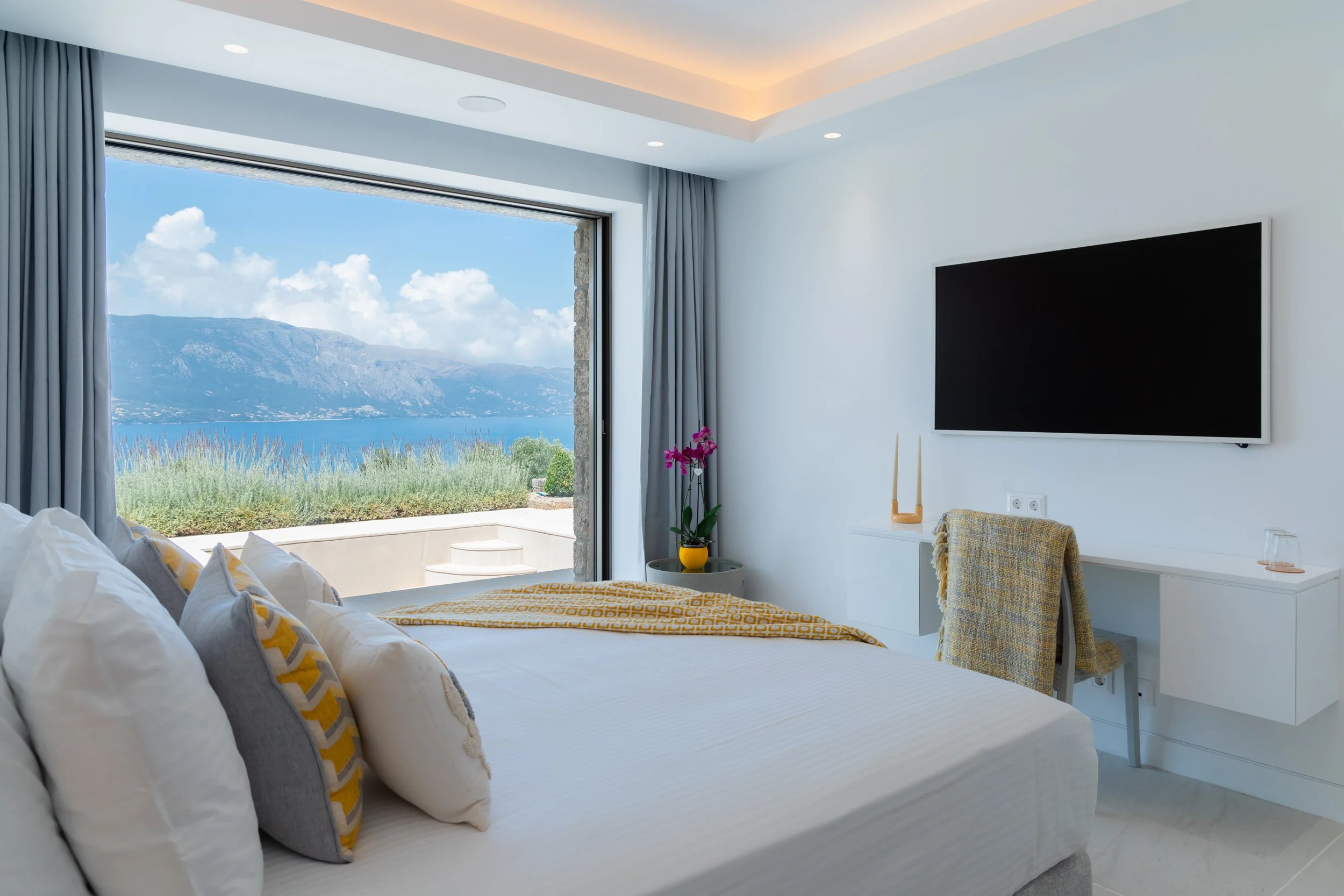 A modern bedroom with a large bed, colorful pillows, and a view of mountains and water outside a large sliding glass door. The room has white walls, a mounted flat-screen TV, a small table with pink flowers, and a chair with a blanket.