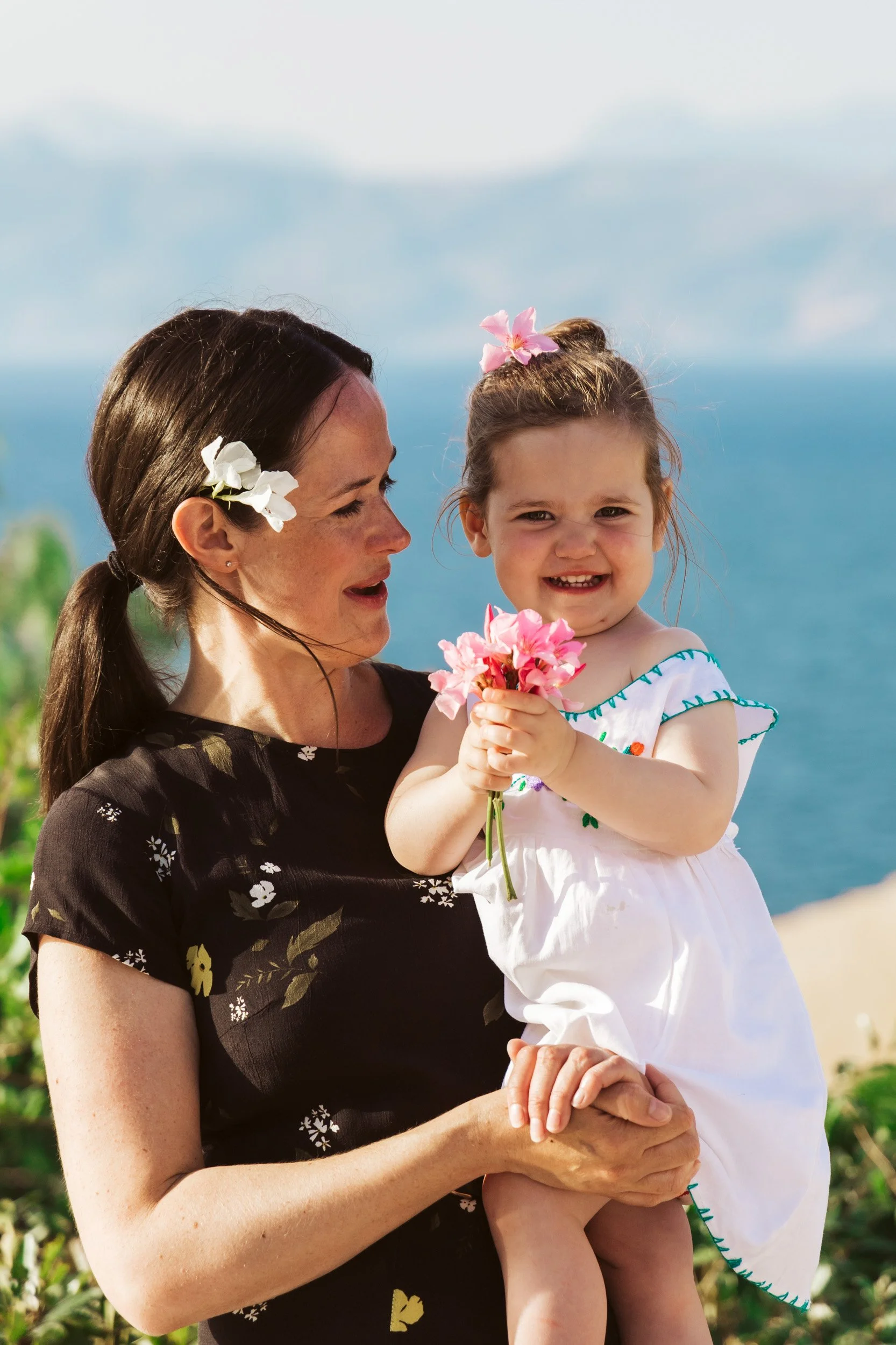 A woman holding a young girl in her arms outdoors near the ocean, both wearing flowers in their hair and smiling, with the girl holding pink flowers.