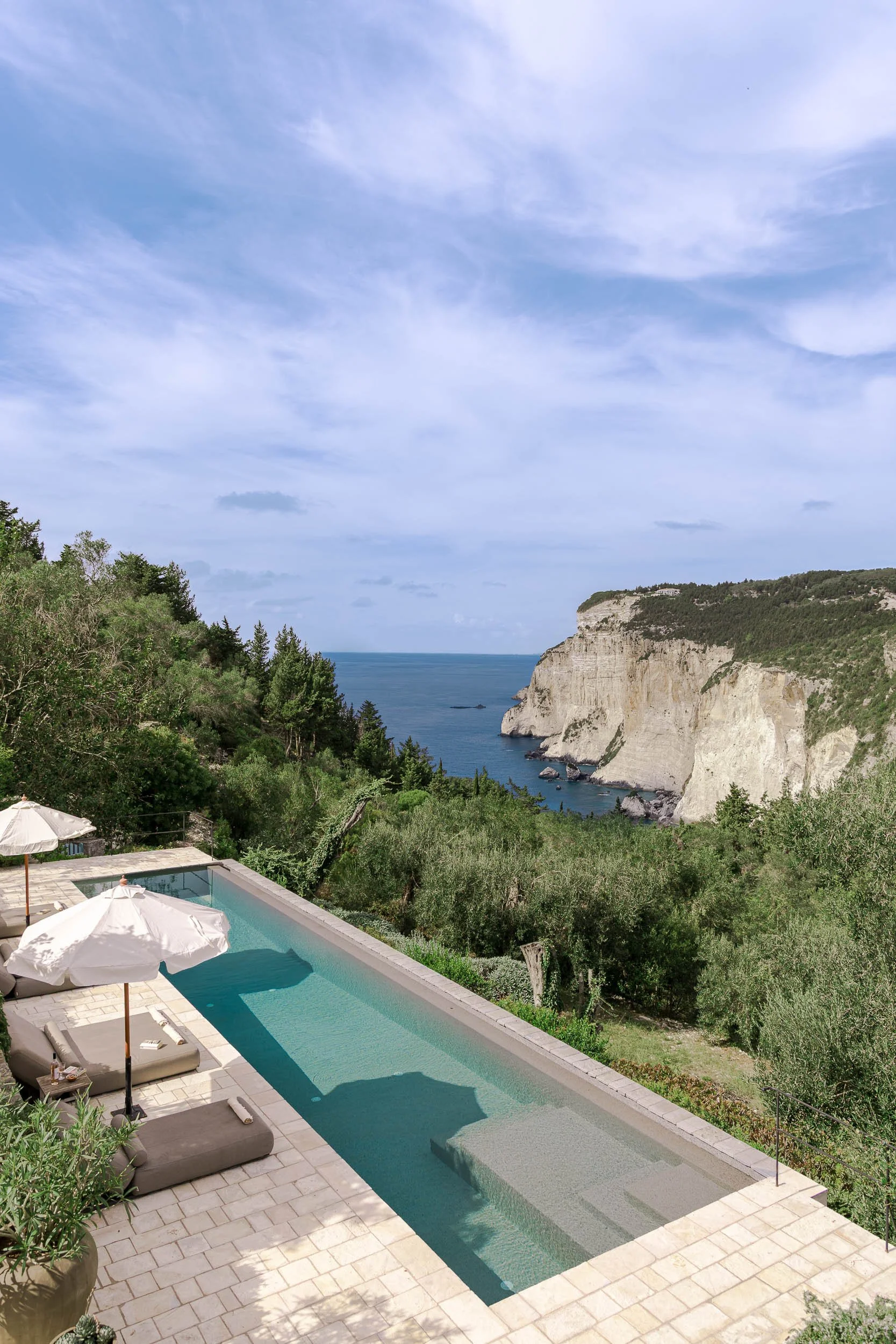 A luxurious rectangular swimming pool with nearby sun loungers and umbrellas, overlooking lush greenery, cliffs, and the ocean under a partly cloudy sky