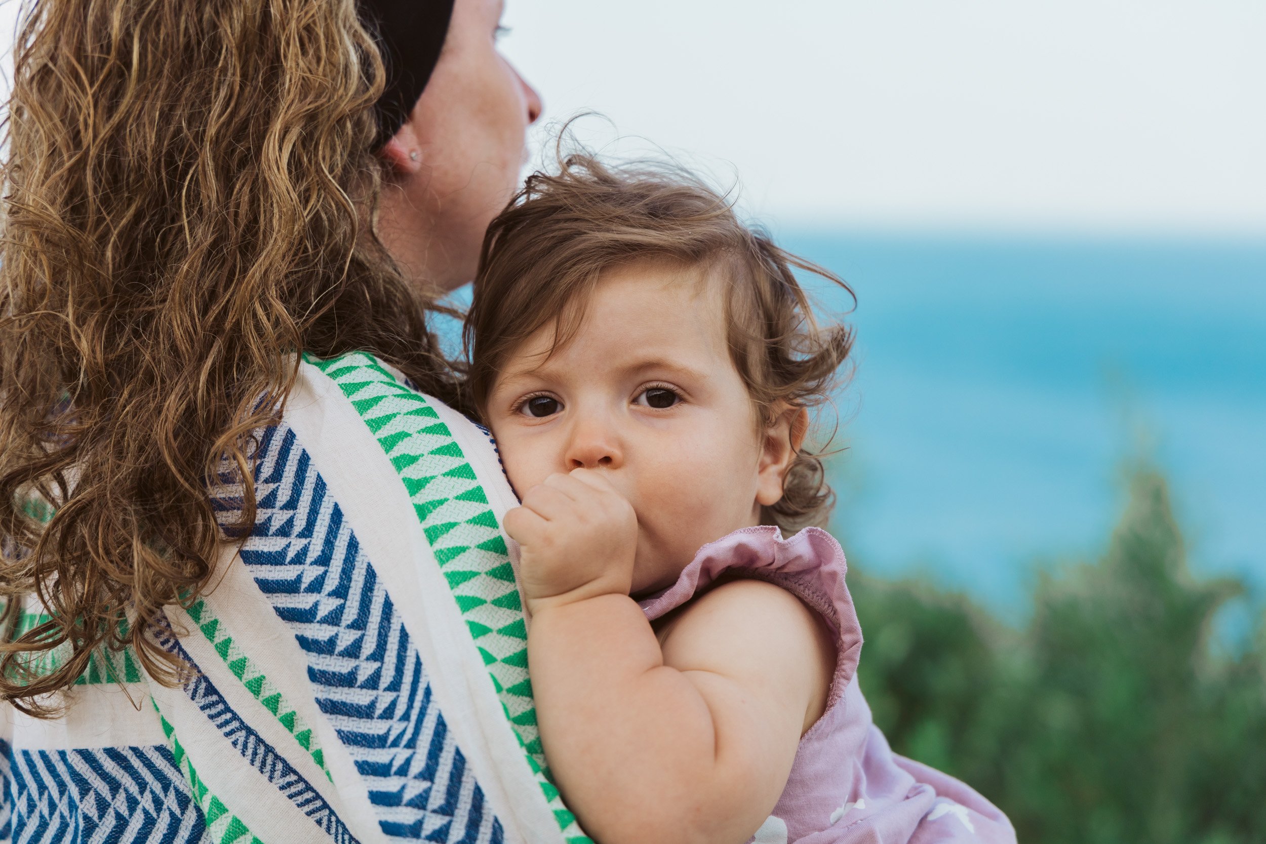 A woman holding a young girl with curly brown hair and big eyes, in front of a blurred outdoor background with trees and water.