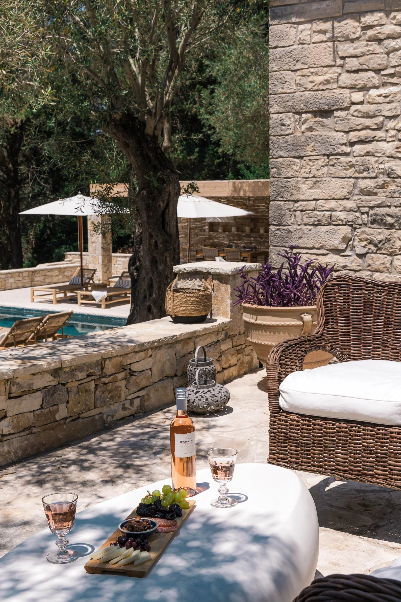 Outdoor patio with wicker armchair, low table with wine, grapes, cheese, and snacks, stone wall, trees, umbrellas, and pool in background.