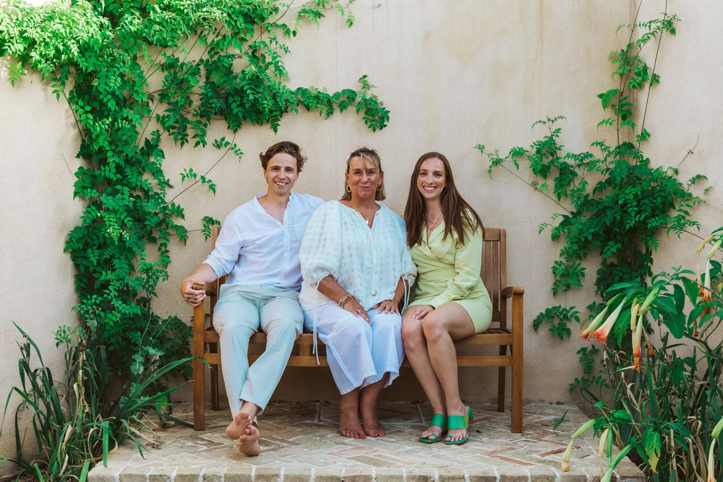Three people sitting on a wooden bench in a garden with green climbing plants on a beige wall behind them. They are smiling and dressed in light summer clothing.