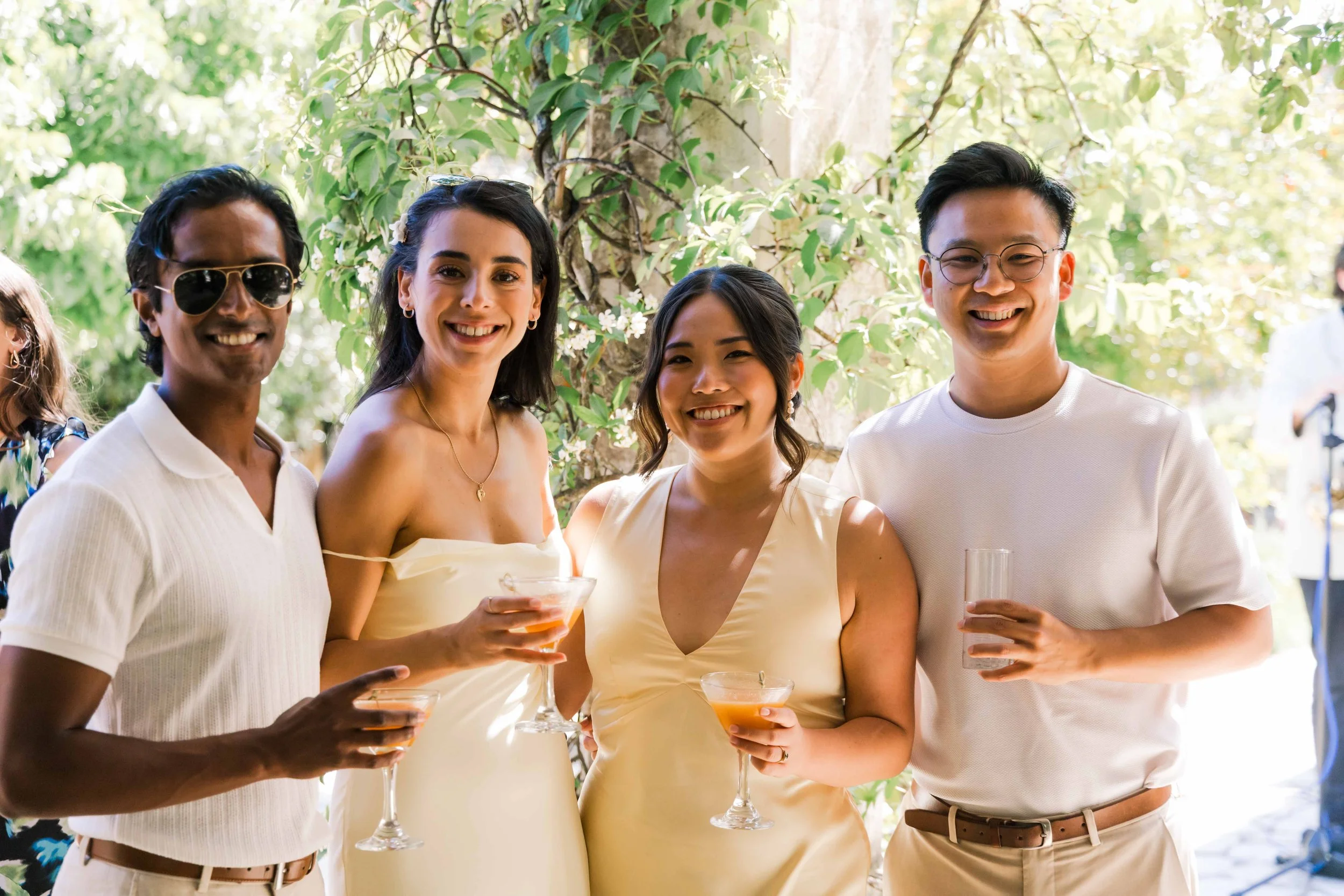 Group of four people smiling and holding drinks at an outdoor gathering surrounded by greenery.