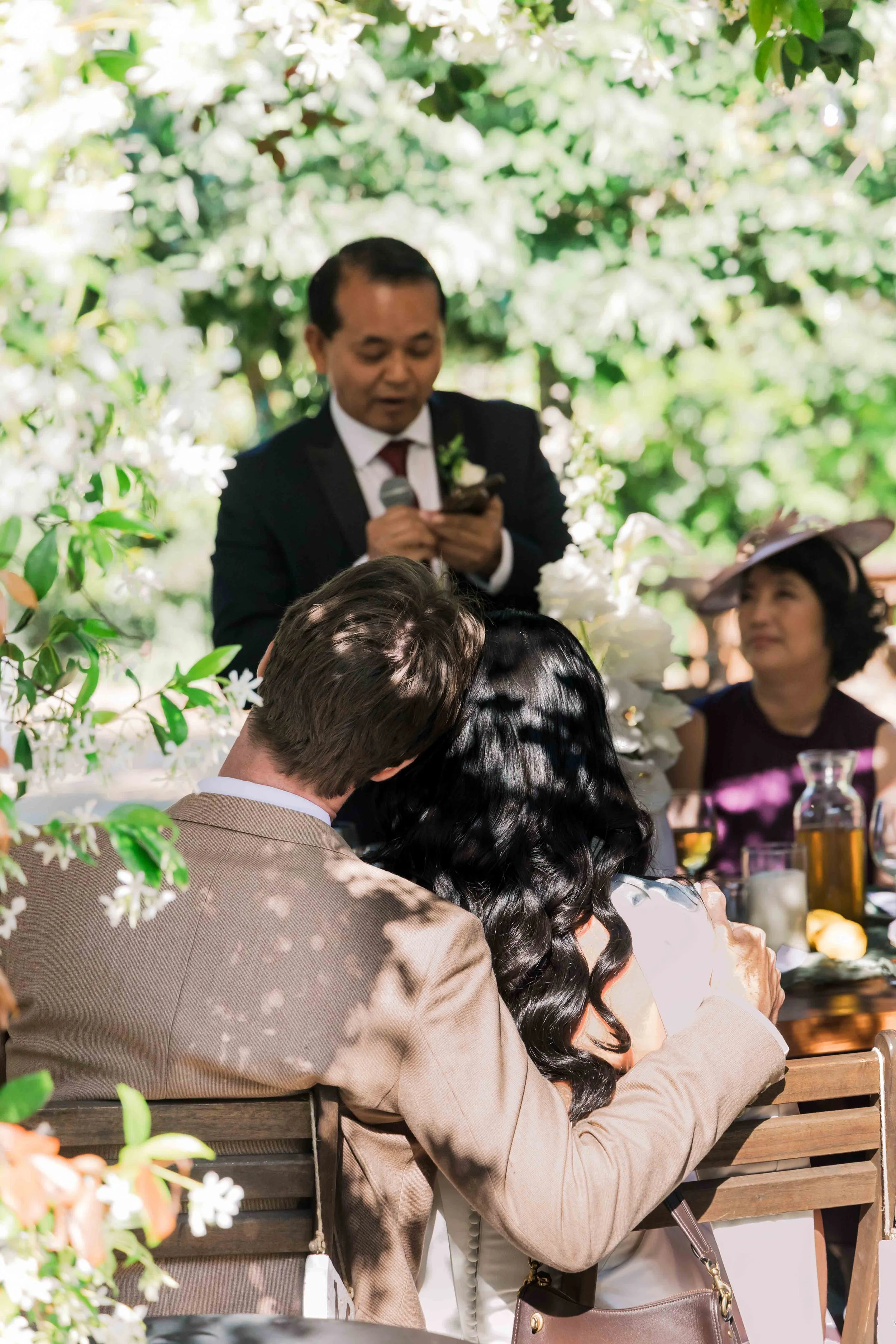 A wedding ceremony outdoors with a man in a suit giving a speech, a couple sitting with their heads together, an older woman in a hat sitting at the table, and lush greenery in the background.